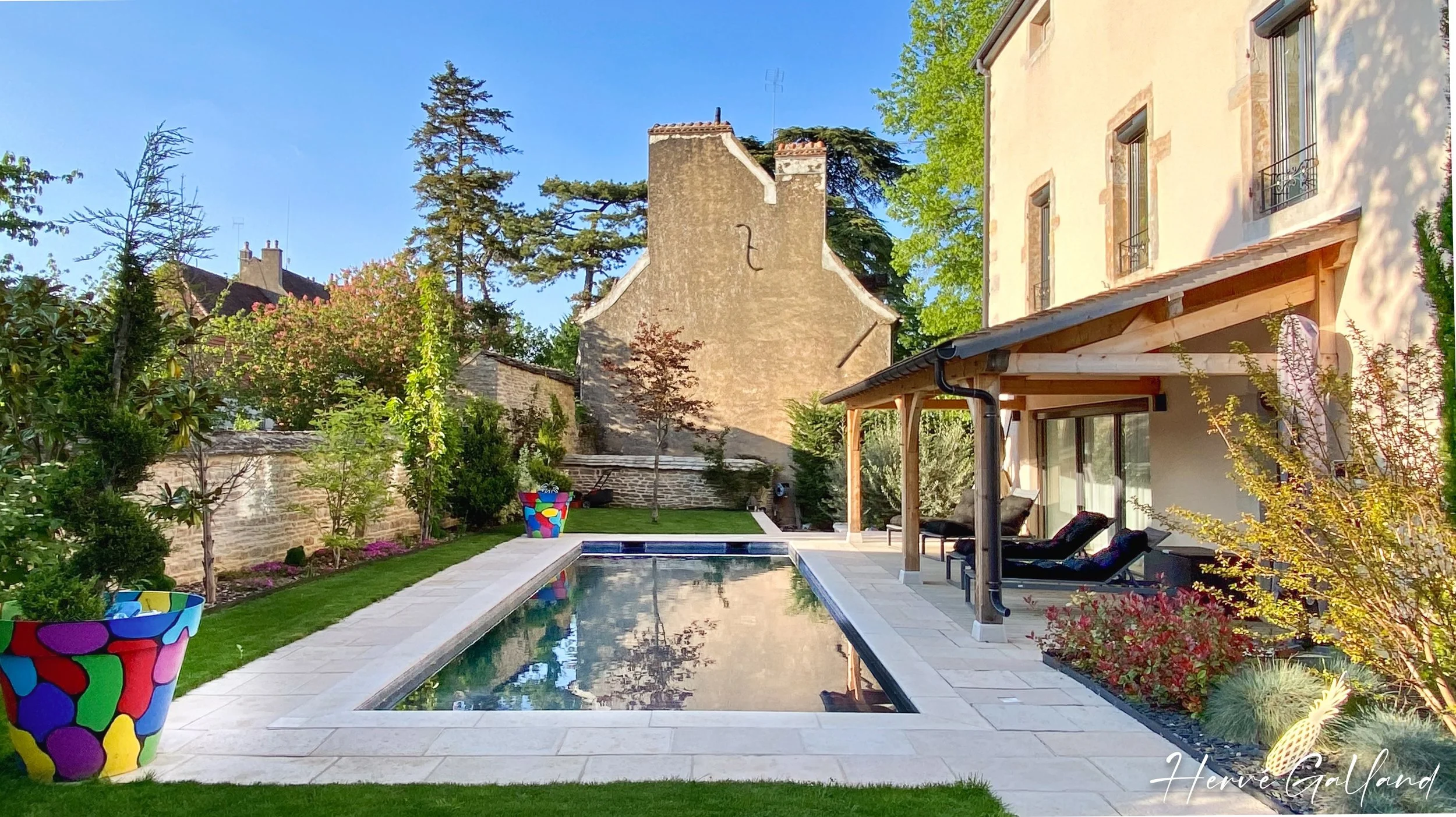 Jardin avec piscine, plantes vertes, maison ancienne en pierre, parasol en bois avec fauteuils, pots de fleurs colorés, ciel bleu.