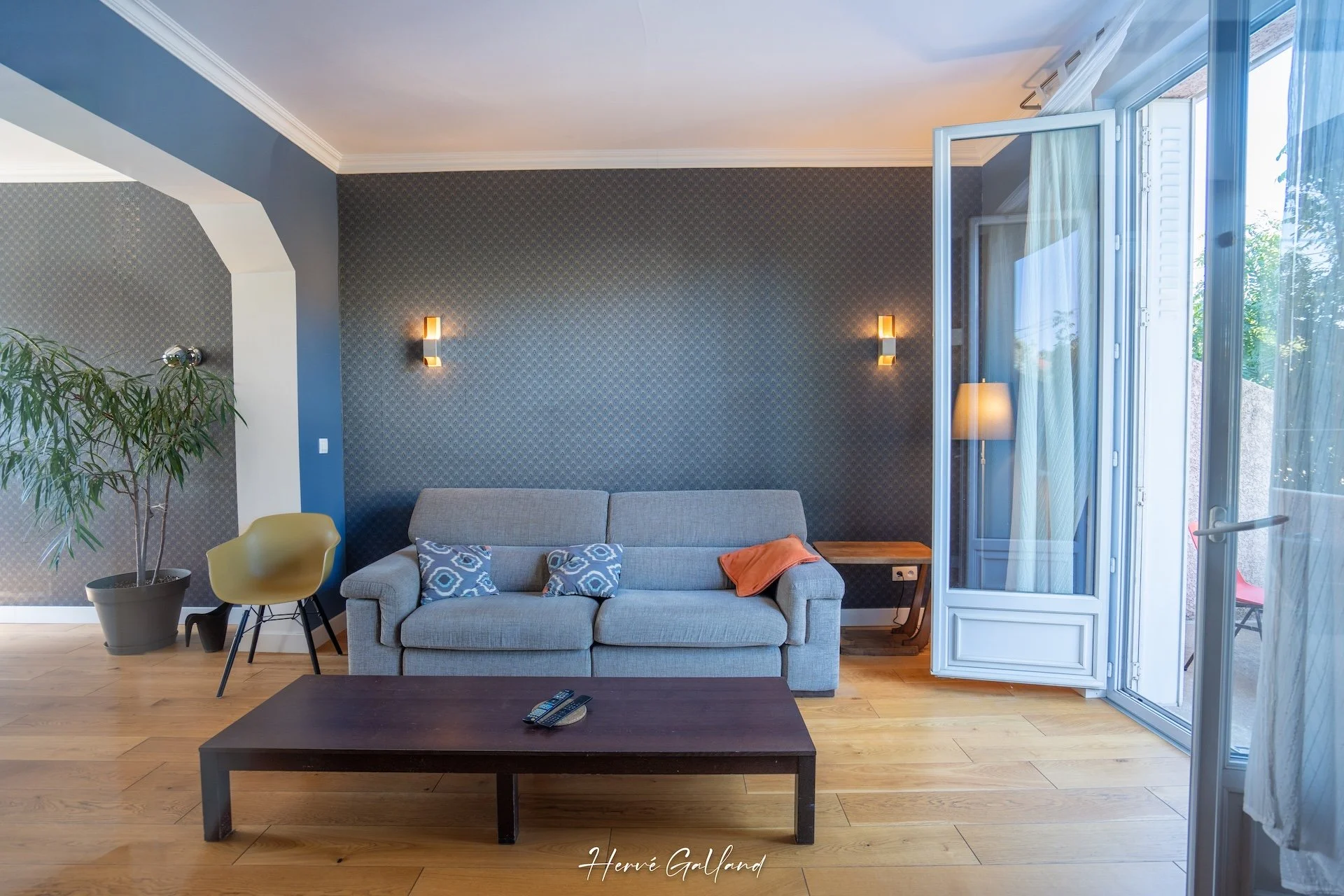 Salon with gray sofa, wooden coffee table, potted plant, yellow chair, patterned wall, open glass door, curtains, side table with lamp.