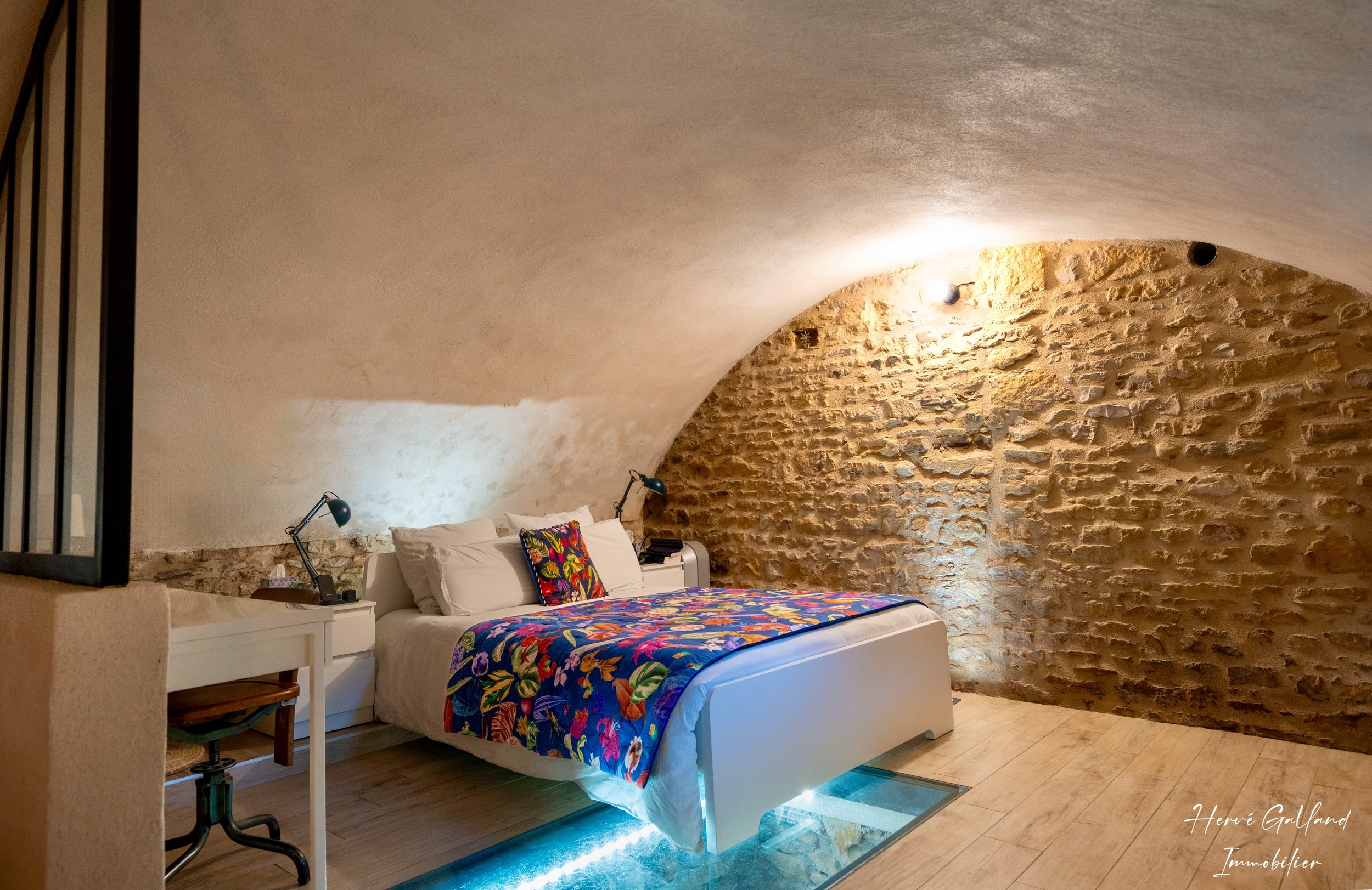 Chambre moderne avec un lit au drapeau floral coloré, murs en pierre apparente et plafond voûté, éclairage tamisé.