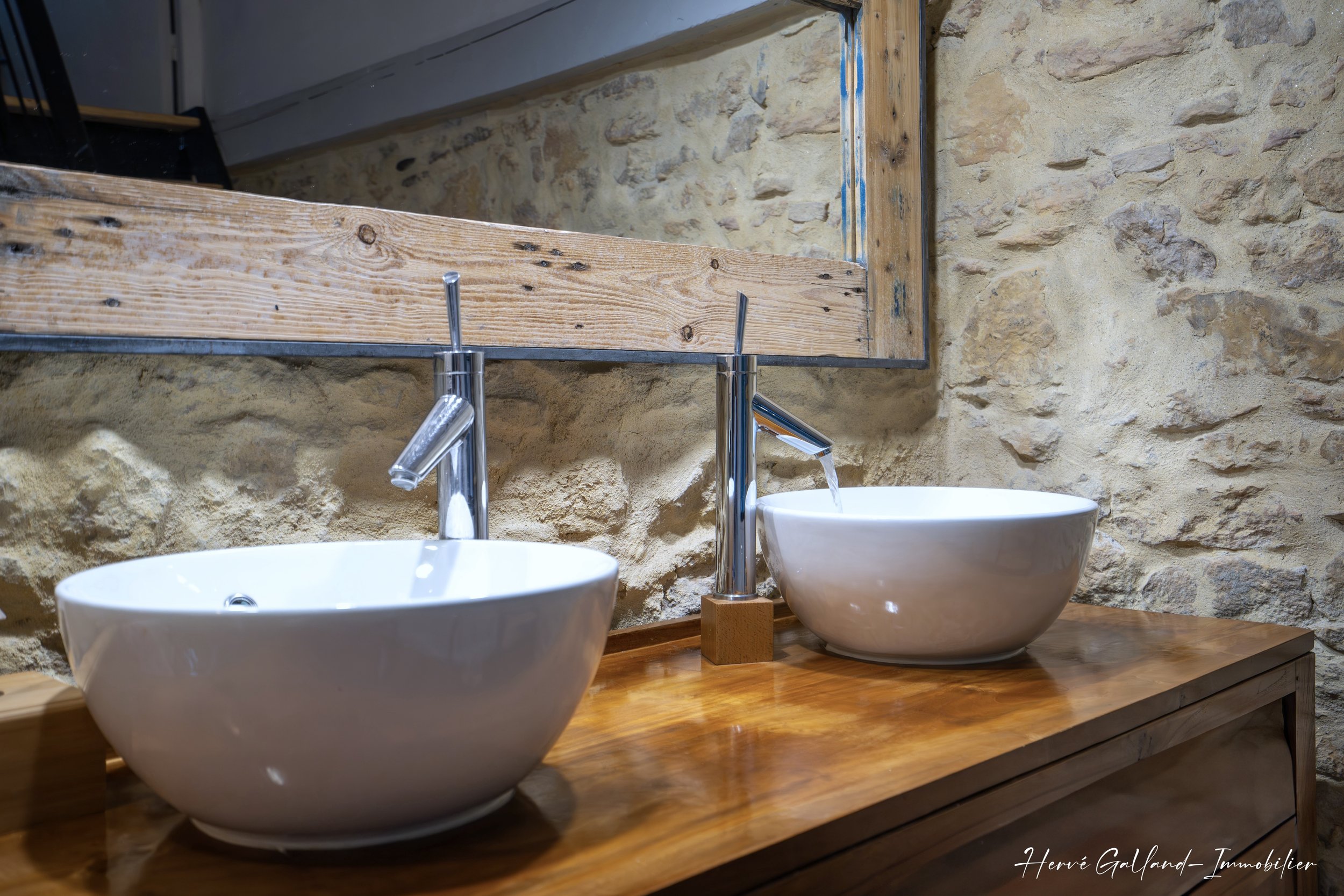 Deux lavabos ronds blancs avec robinets en chrome sur une surface en bois, dans une salle de bain avec murs en pierre et un miroir au-dessus.