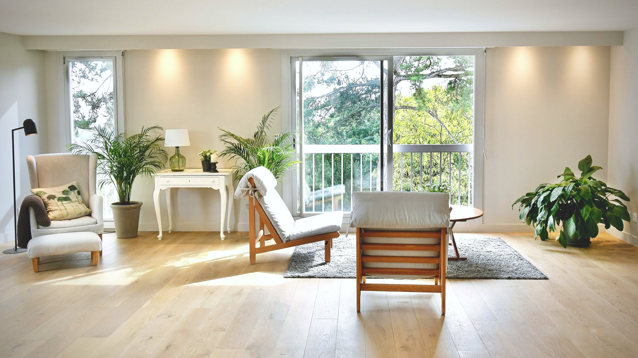 Salon lumineux avec grande fenêtre donnant sur un balcon entouré d'arbres, comprenant un fauteuil beige, un fauteuil en bois avec coussin blanc, une petite table ronde en bois, plusieurs plantes vertes, une lampe de table blanche et un tapis gris.