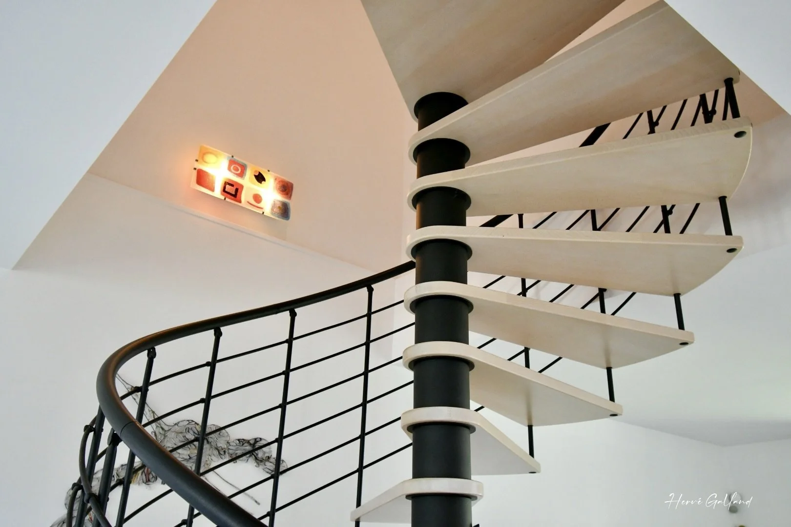 Escalier spiral moderne en bois clair avec main courante en métal noir dans un espace intérieur lumineux.