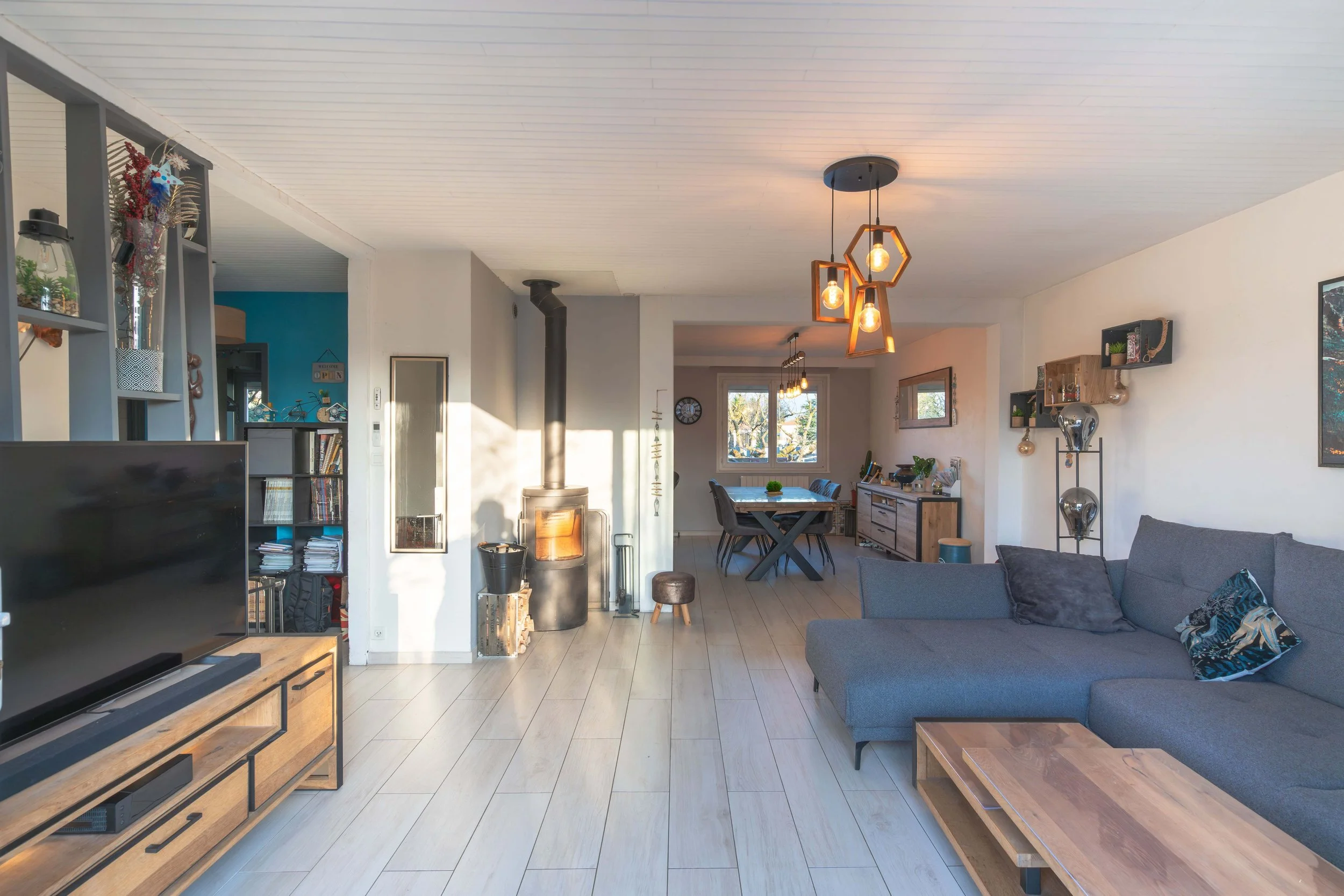 Salon spacieux avec canapé gris, cheminée noire, télévision sur meuble en bois, table basse en bois, murs blancs, étagères avec décorations, et salle à manger au fond avec grande fenêtre