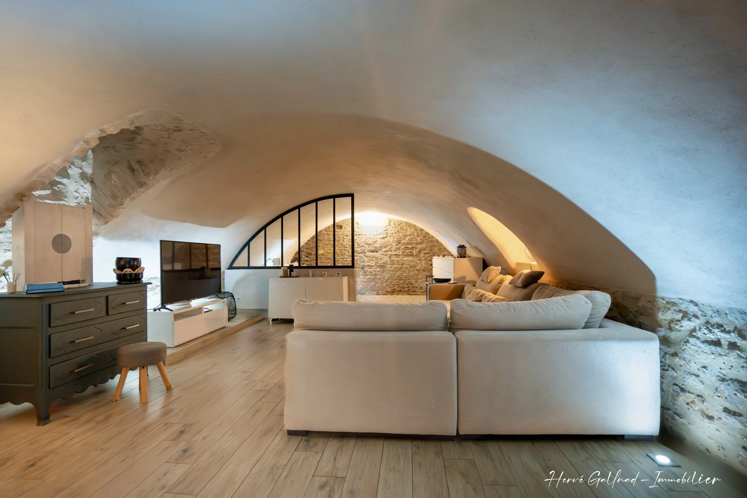 Salon avec canapé beige, télévision, meuble de rangement, et plafond en voûte en pierre dans une pièce lumineuse.