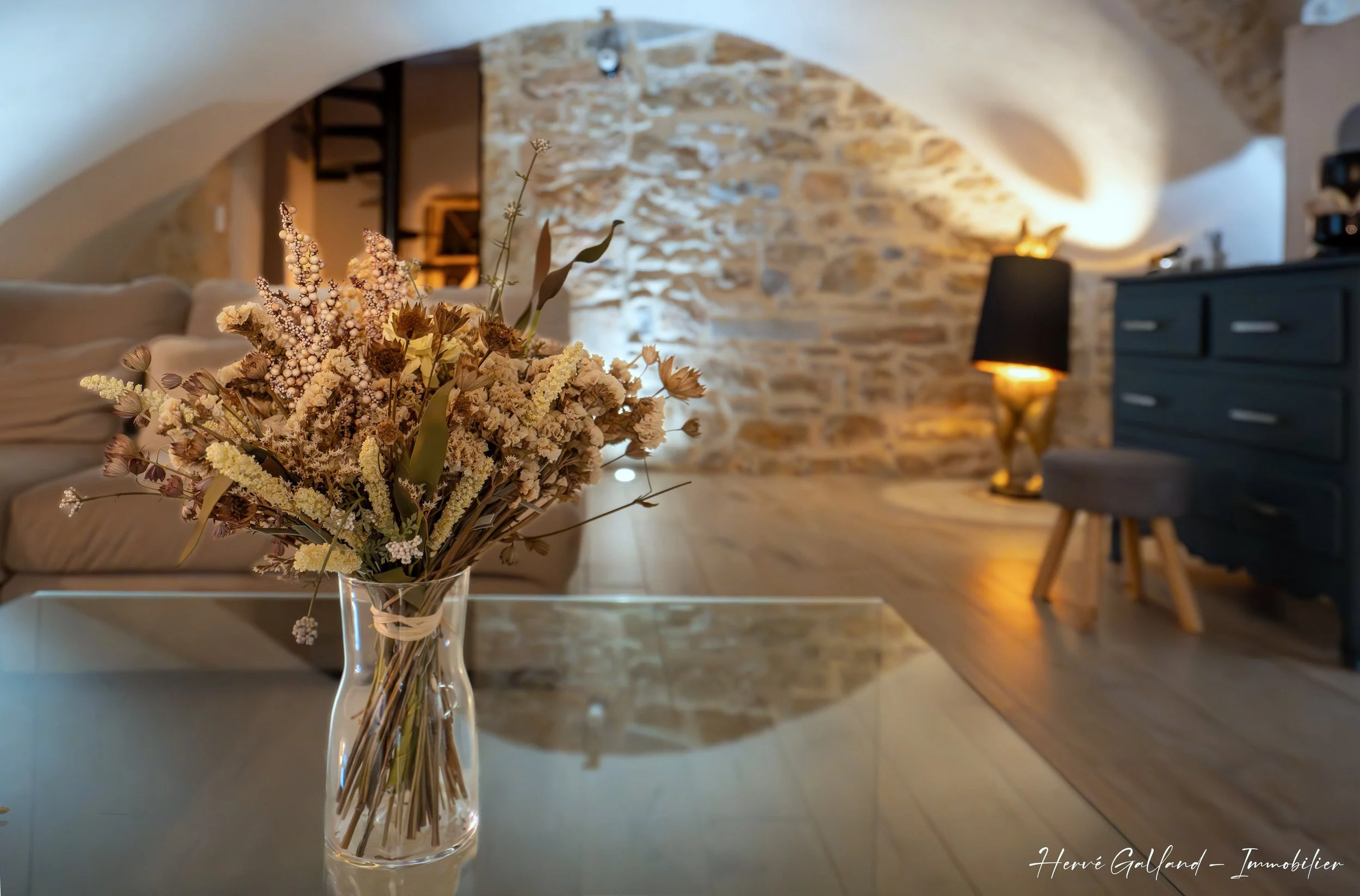 Bouquet de fleurs séchées dans un vase en verre sur une table en verre dans un salon à l'ambiance chaleureuse avec un mur en pierre, une lampe et des meubles en bois.