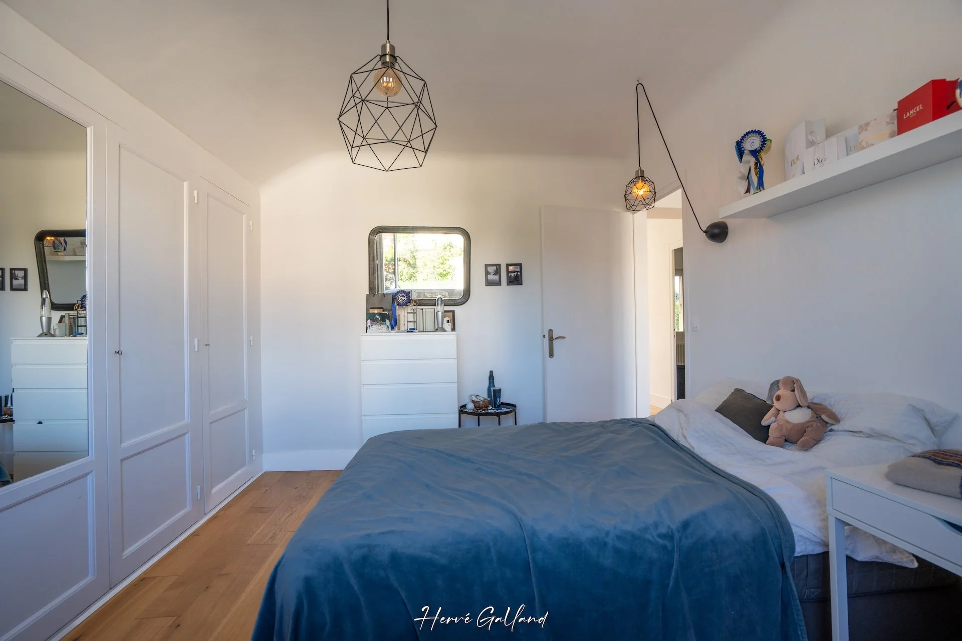 Chambre avec un lit, une armoire avec miroir, des lampes suspendues, une bibliothèque, un miroir, et un petit meuble avec des objets, ambiance lumineuse et décoration simple.