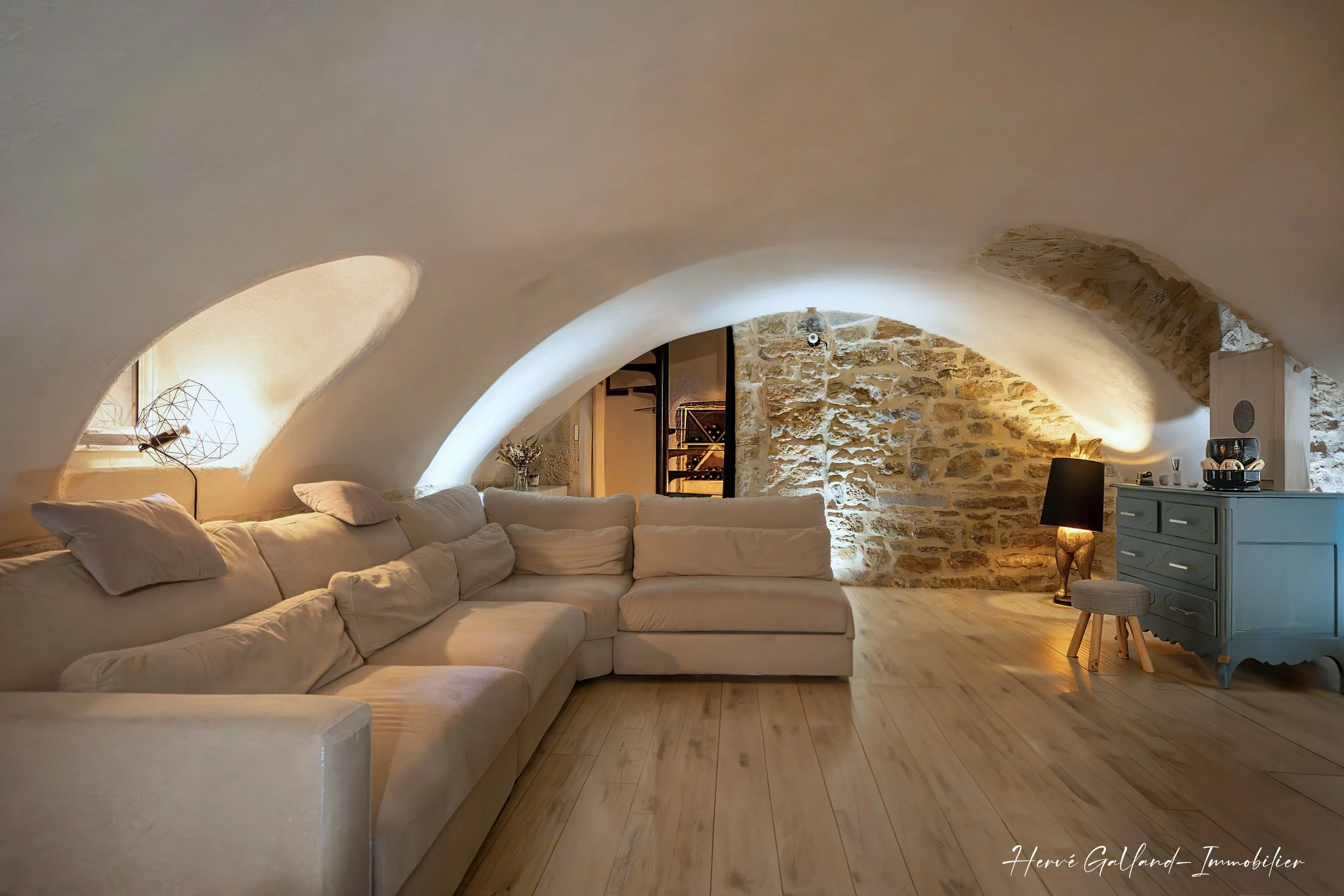 Salon dans une pièce avec plafond en arche, canapé beige, murs en pierre, lampes sur table, sol en bois.