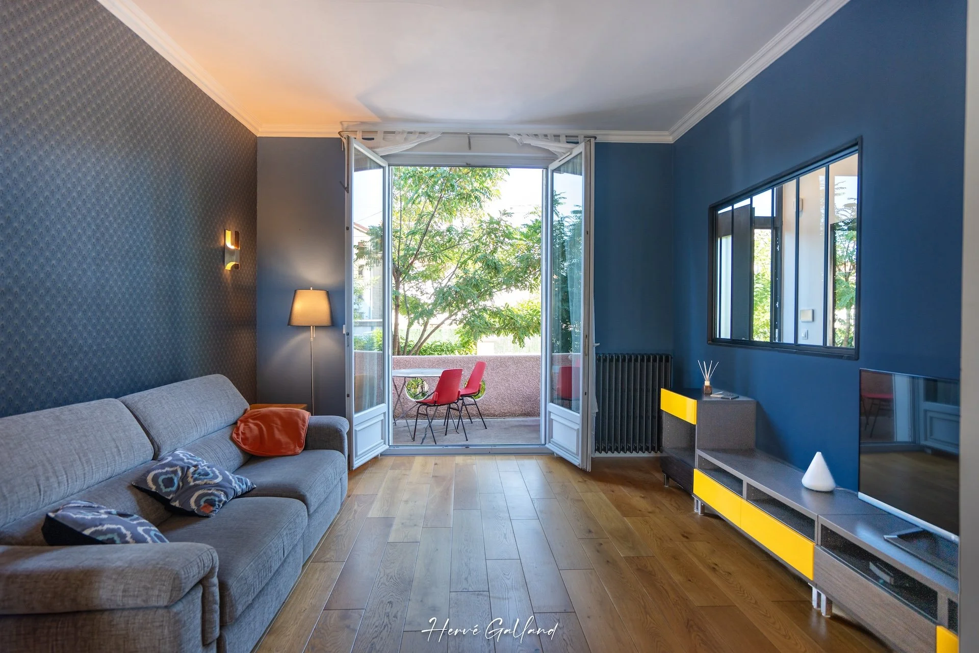 Salon lumineux avec mur bleu, canapé gris, lampadaire, table sur balcon avec chaises rouges, télévision avec meuble beige et jaune, fenêtre et porte-fenêtre donnant sur balcon avec vue sur arbres.
