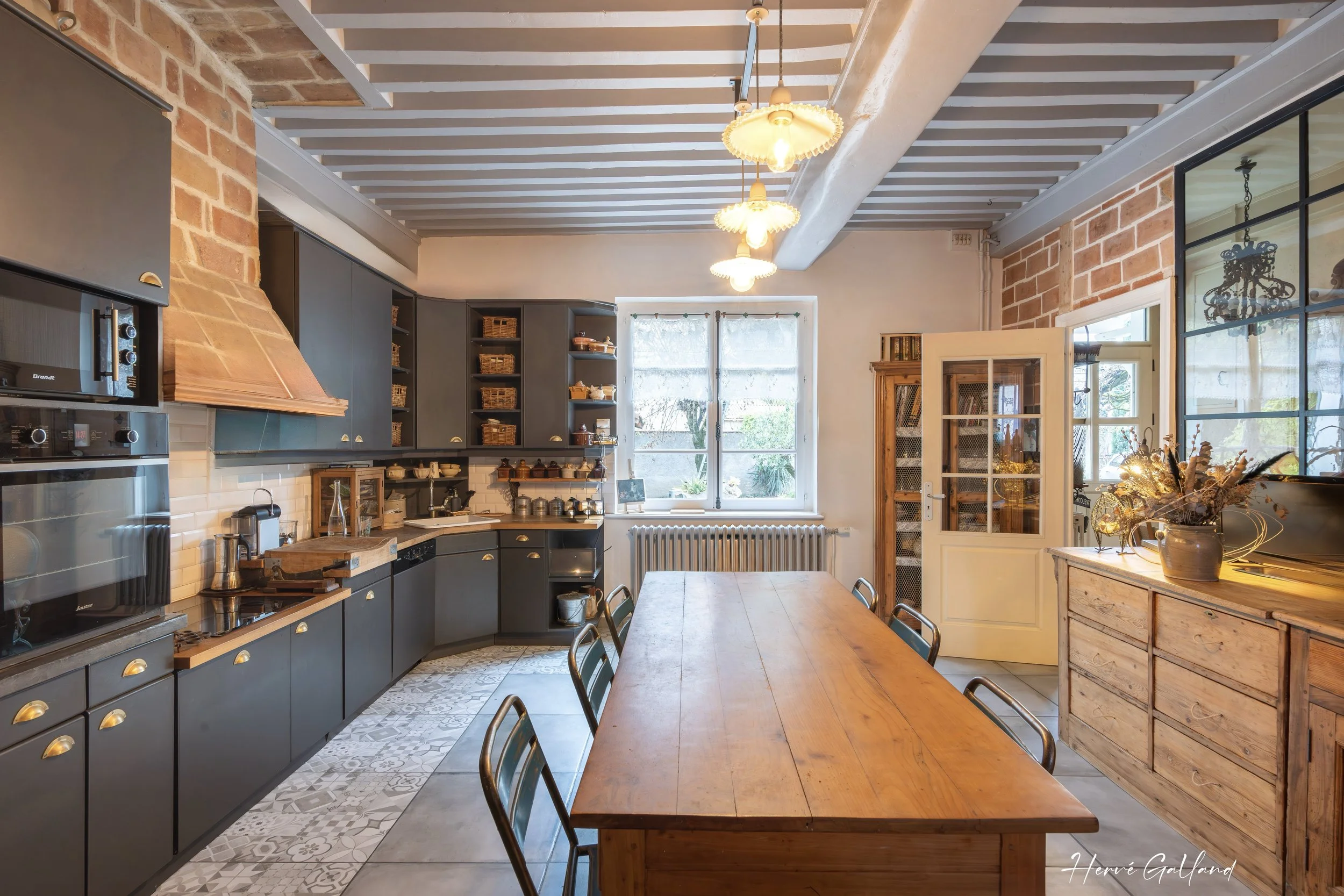 Cuisine industrielle avec mur en briques, meubles en bois et équipements modernes, grande table en bois au centre, luminaires vintage au plafond, fenêtre laissant entrer la lumière naturelle, décoration chaleureuse.
