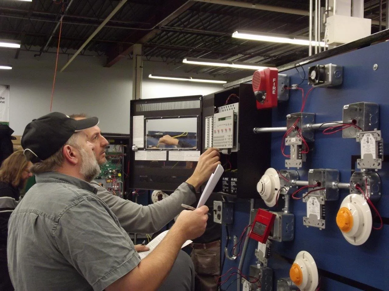 A man with a beard and wearing a baseball cap working on electronic and electrical equipment mounted on a blue panel, with a notebook in his hand, in a workshop or industrial setting.