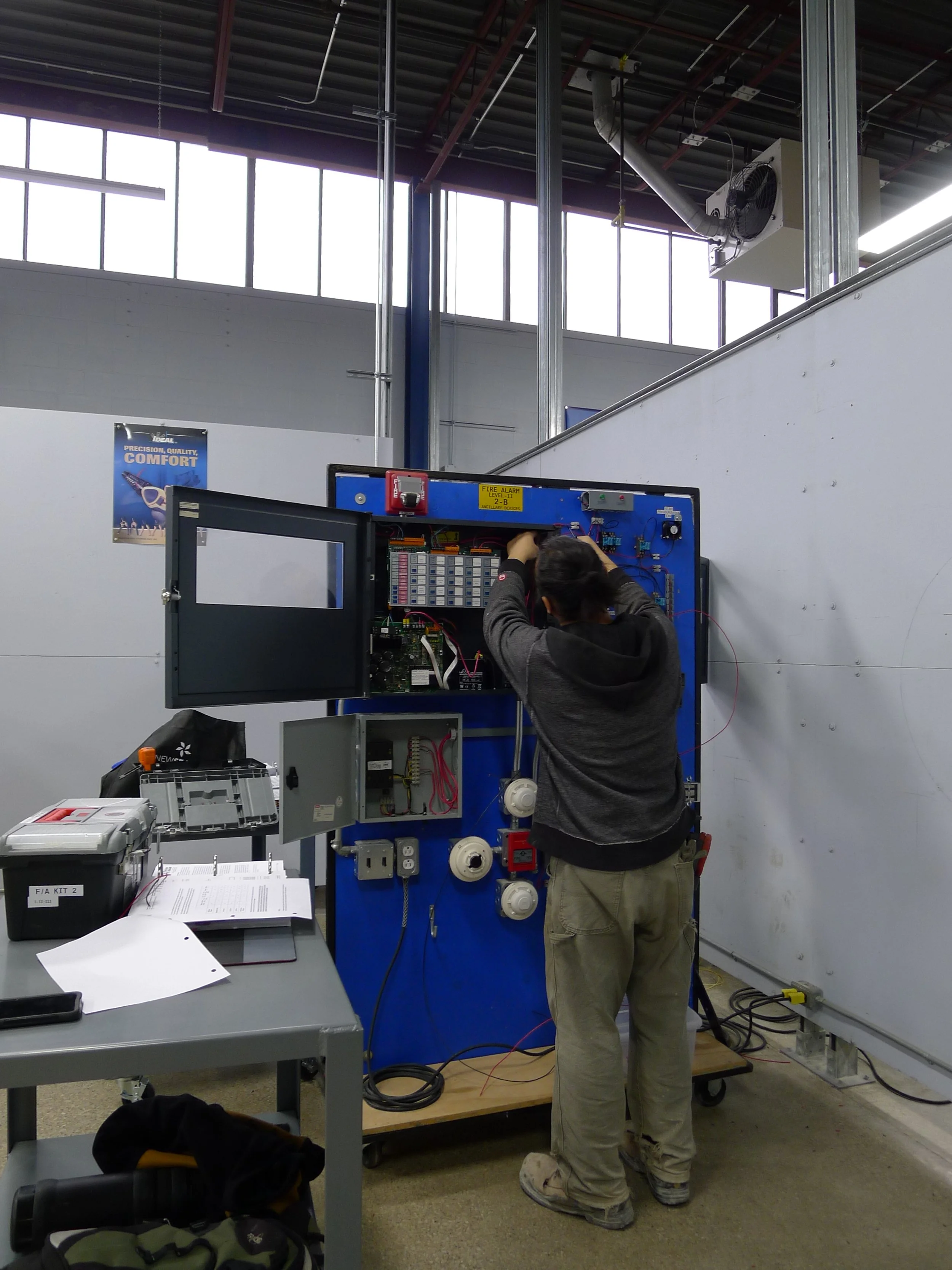 A person working on an electrical panel in an industrial or training setting with tools and documents on a nearby table.