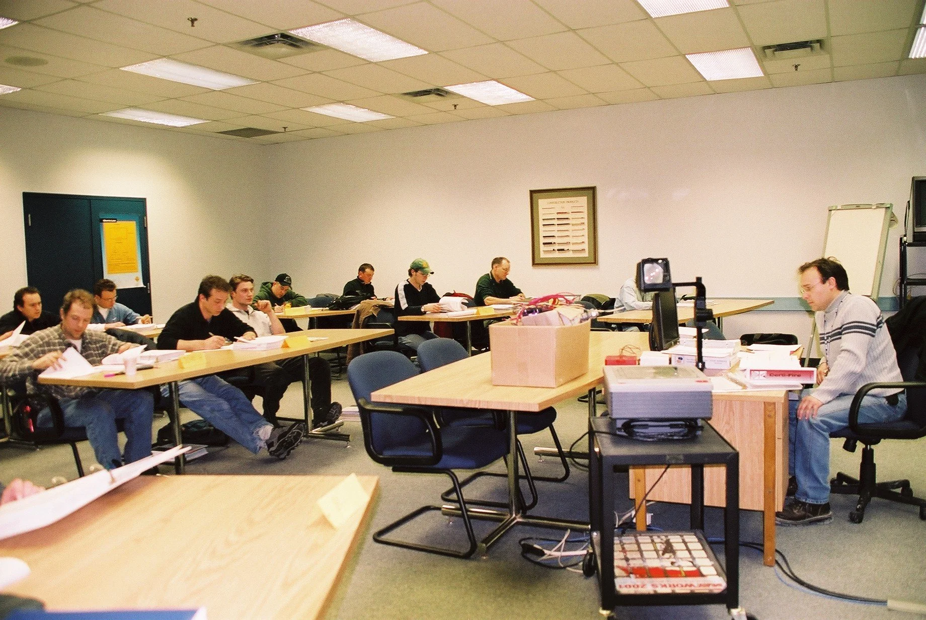 A classroom with students sitting at desks, working on papers, with a teacher at the front near a whiteboard and a desk with books and supplies.