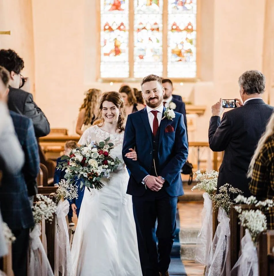 A bride and groom walking down the aisle in a church, with the bride holding a bouquet of white, red, and purple flowers, and the groom dressed in a blue suit. Guests are taking photos, and stained glass windows are visible in the background.