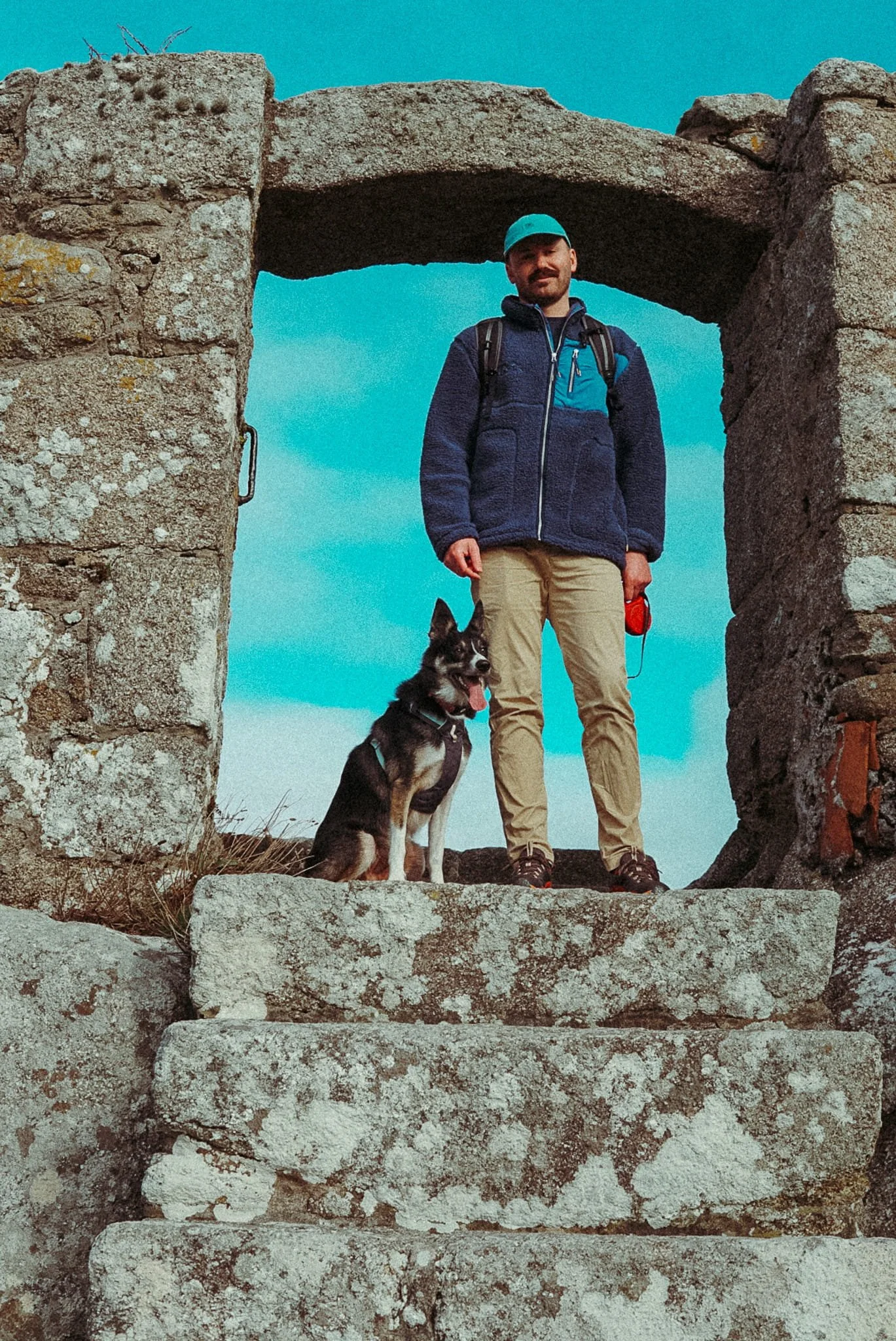 A man with a backpack and a hat standing on stone steps with a dog at his side, framed by ancient stone ruins under a blue sky.