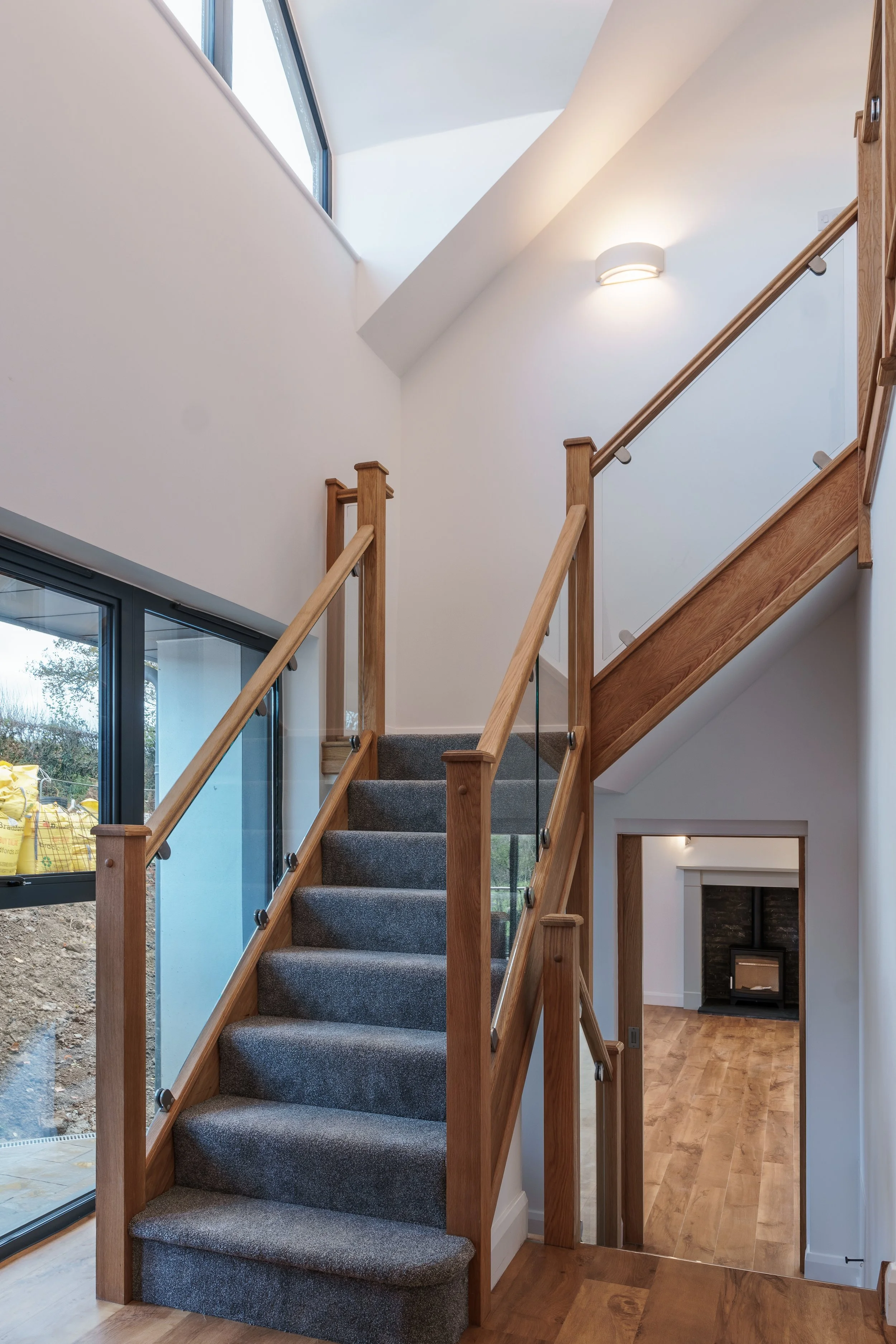 Interior view of a modern staircase with wooden railings, gray carpeted steps, and a large window letting in natural light, leading to a mezzanine or upper floor in a house.