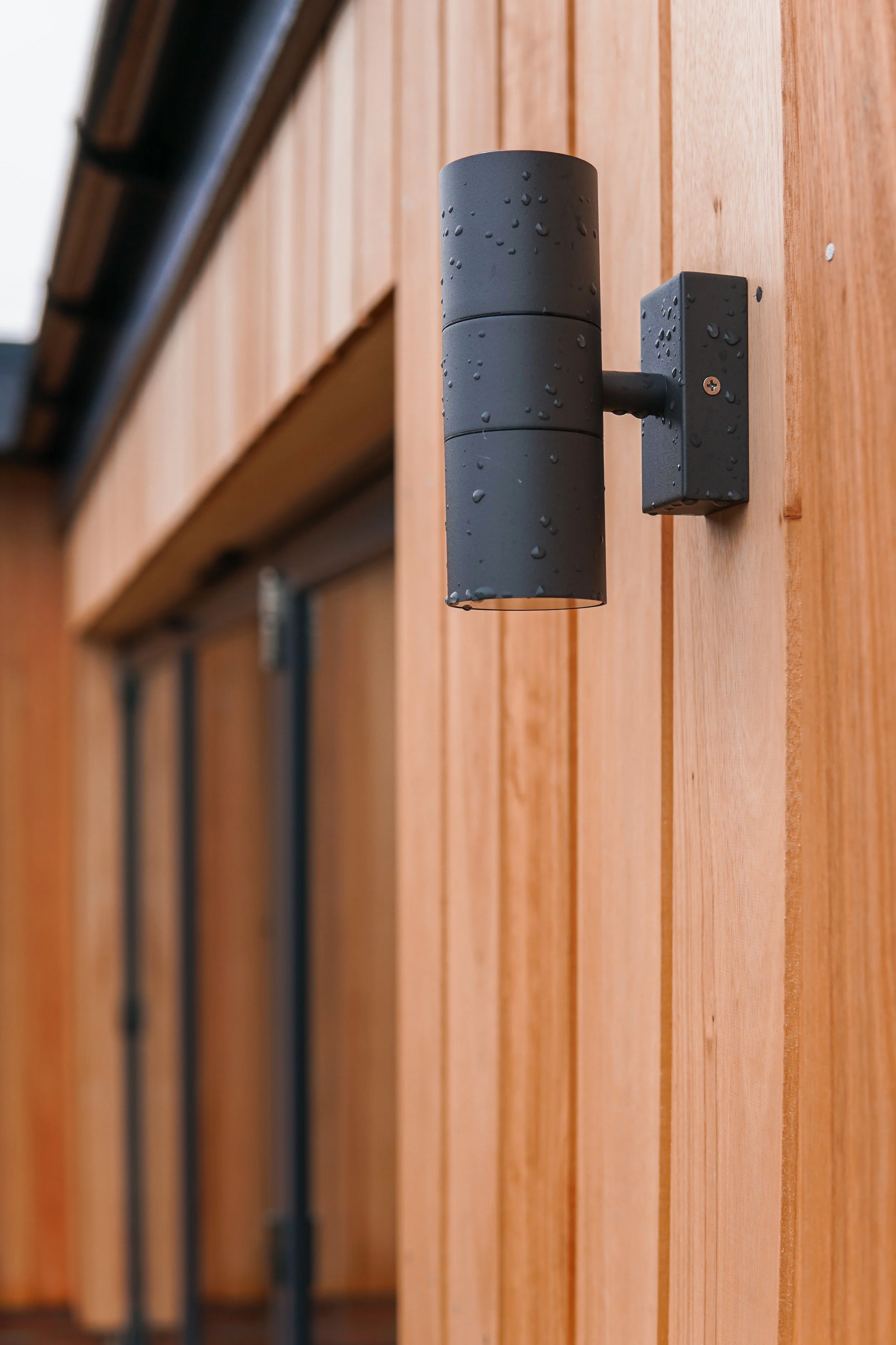 Close-up of a black outdoor wall light mounted on a wooden wall with water droplets on its surface.