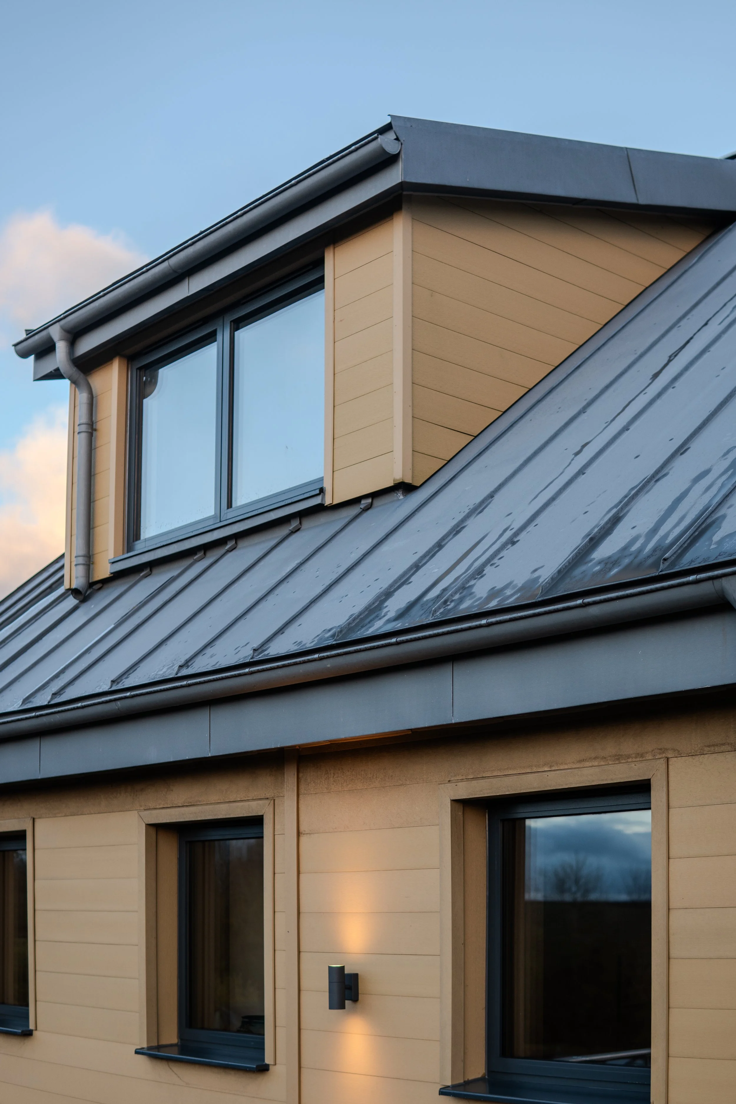 Close-up of a modern house with beige siding, metal roof, and dormer window at sunset.