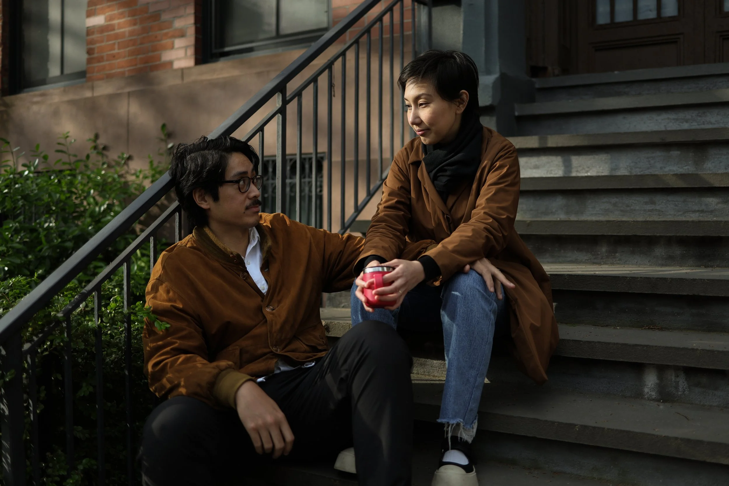 A man and woman sit on outdoor stairs, the woman handing the man a red tumbler, both dressed in brown jackets, with greenery and brick building in the background.