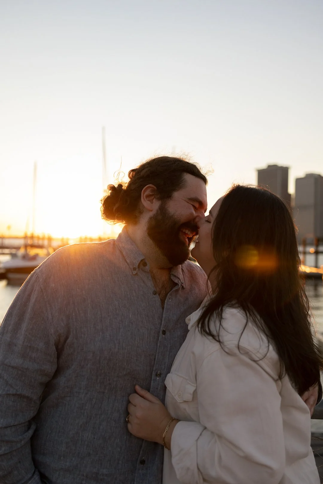 A couple is smiling and about to kiss at sunset near a marina with sailboats and city buildings in the background.
