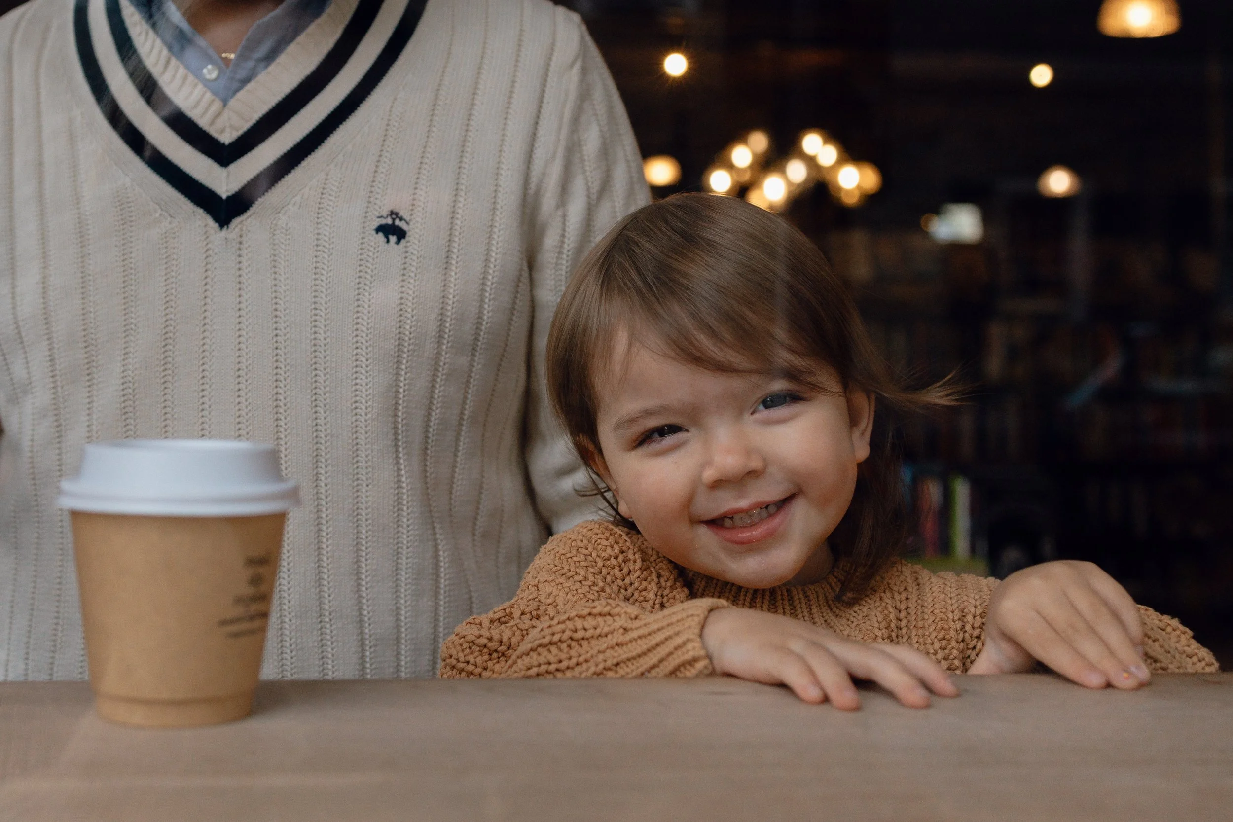 A young girl smiling with her hands on a table, a man standing behind her, and a coffee cup on the table in a cozy indoor setting.