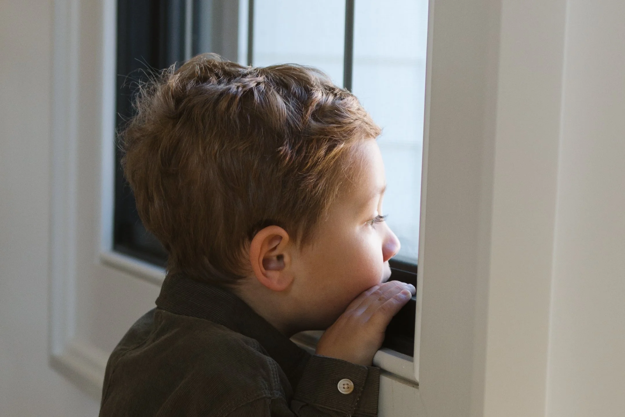 A young boy with short curly hair gazing out of a window, resting his chin on his hands.