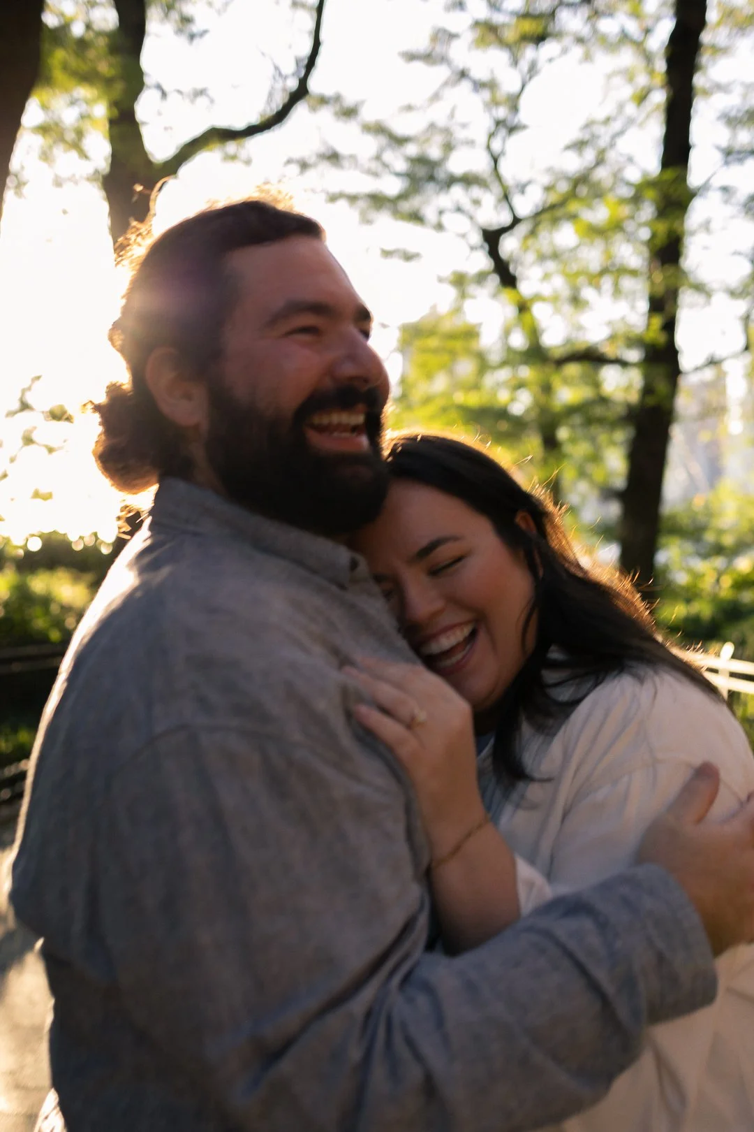 A couple is embracing outdoors with sunlight filtering through trees in the background, both smiling and showing affection.