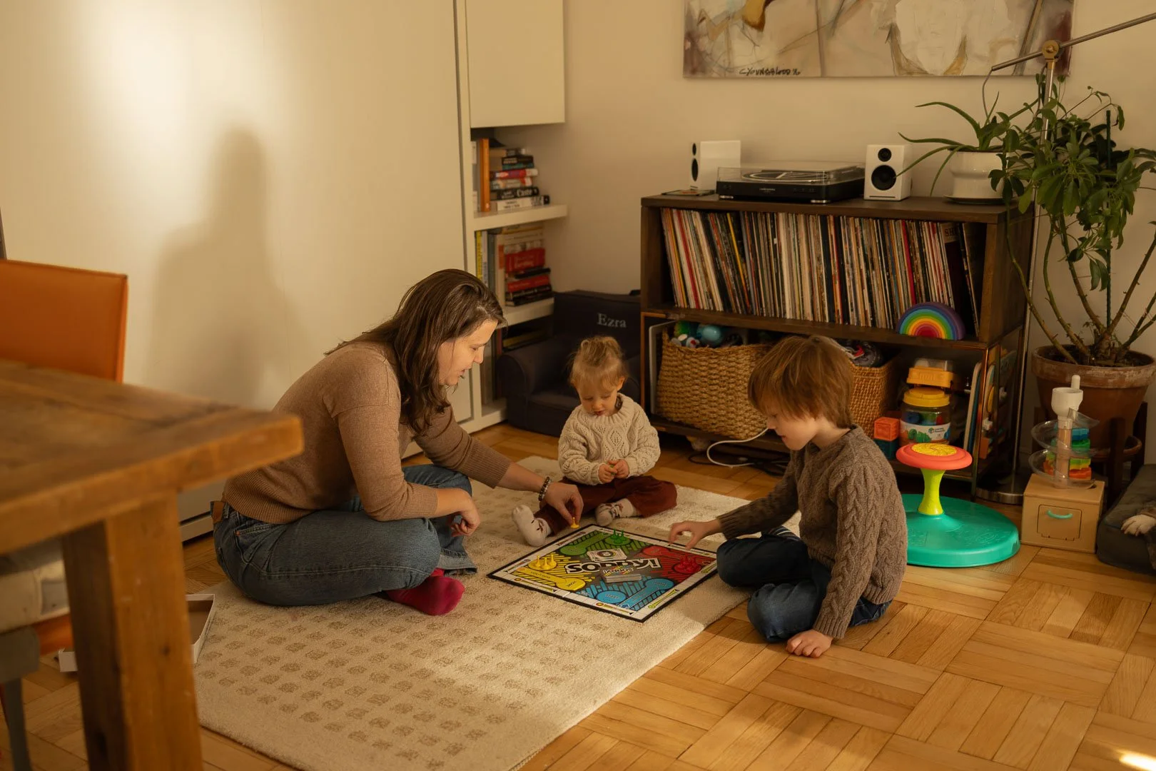 A family playing a board game on the floor in a living room, with a woman and two children surrounded by shelves, books, and toys.