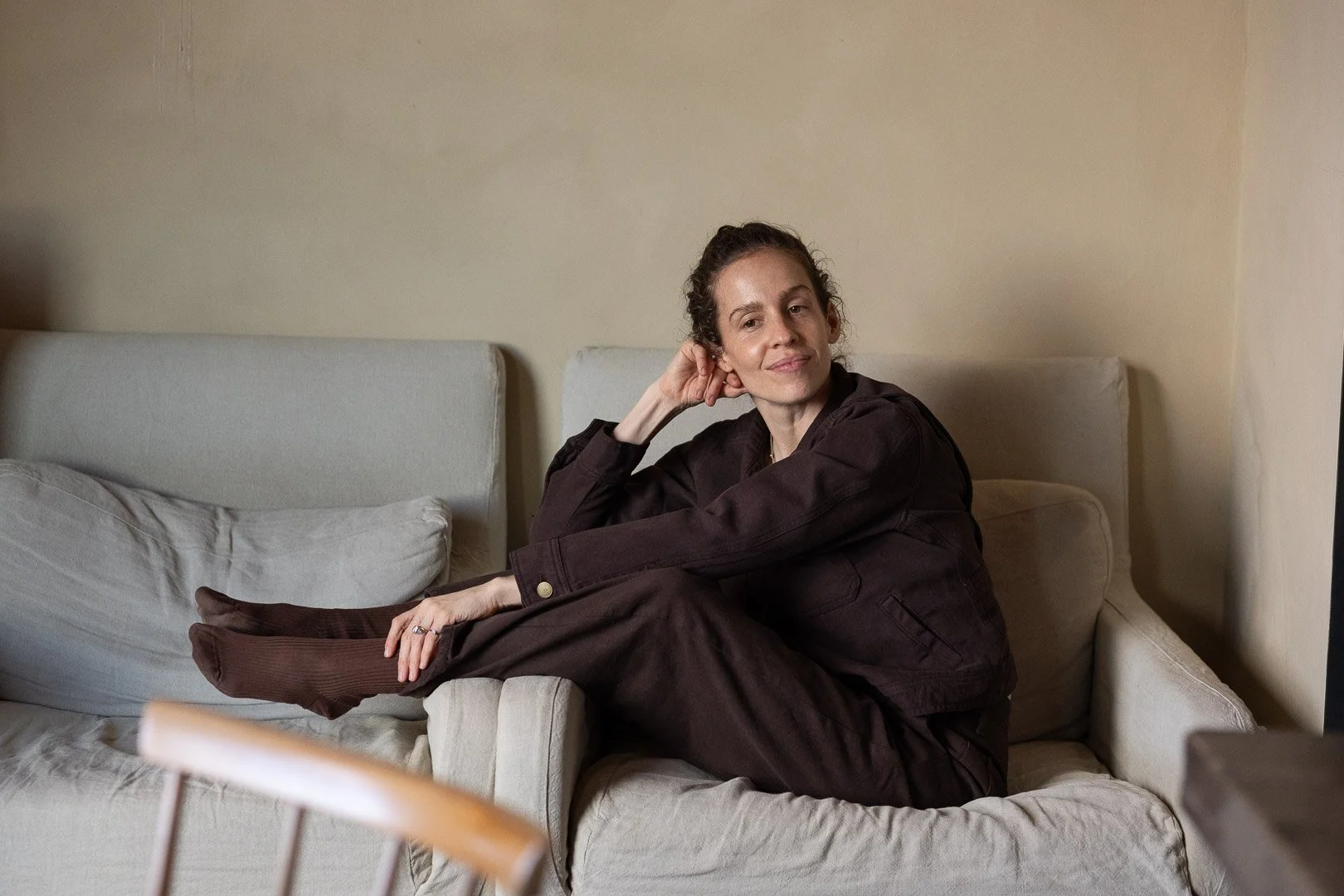A woman with curly hair sitting on a beige sofa, wearing a dark brown jacket and matching pants, with one leg crossed over the other and resting on the sofa, in a room with a plain beige wall.