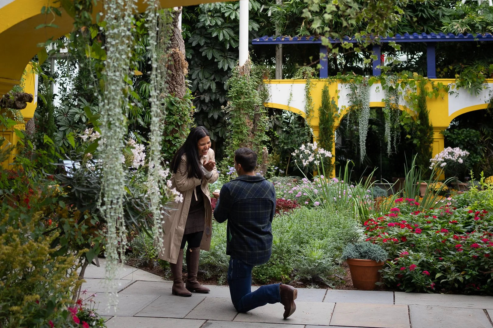 A woman is proposing marriage to a man in a garden filled with colorful flowers, with a yellow and white building in the background.