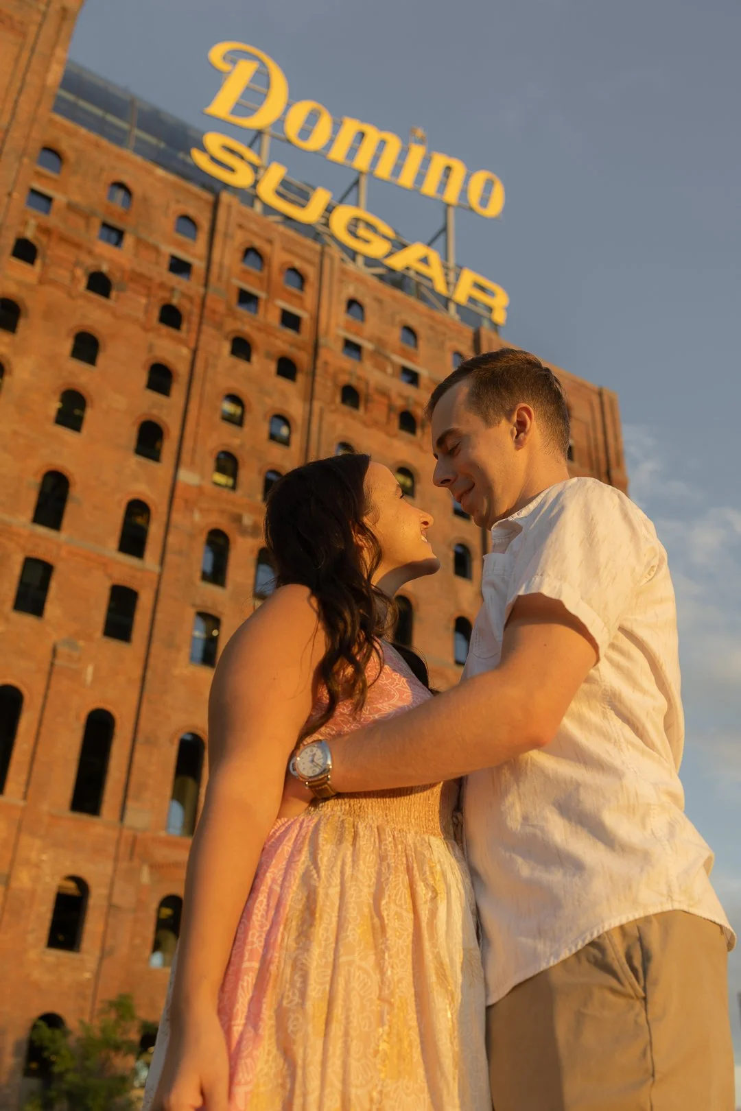 A couple smiling and embracing in front of the Domino Sugar sign and brick building at sunset.