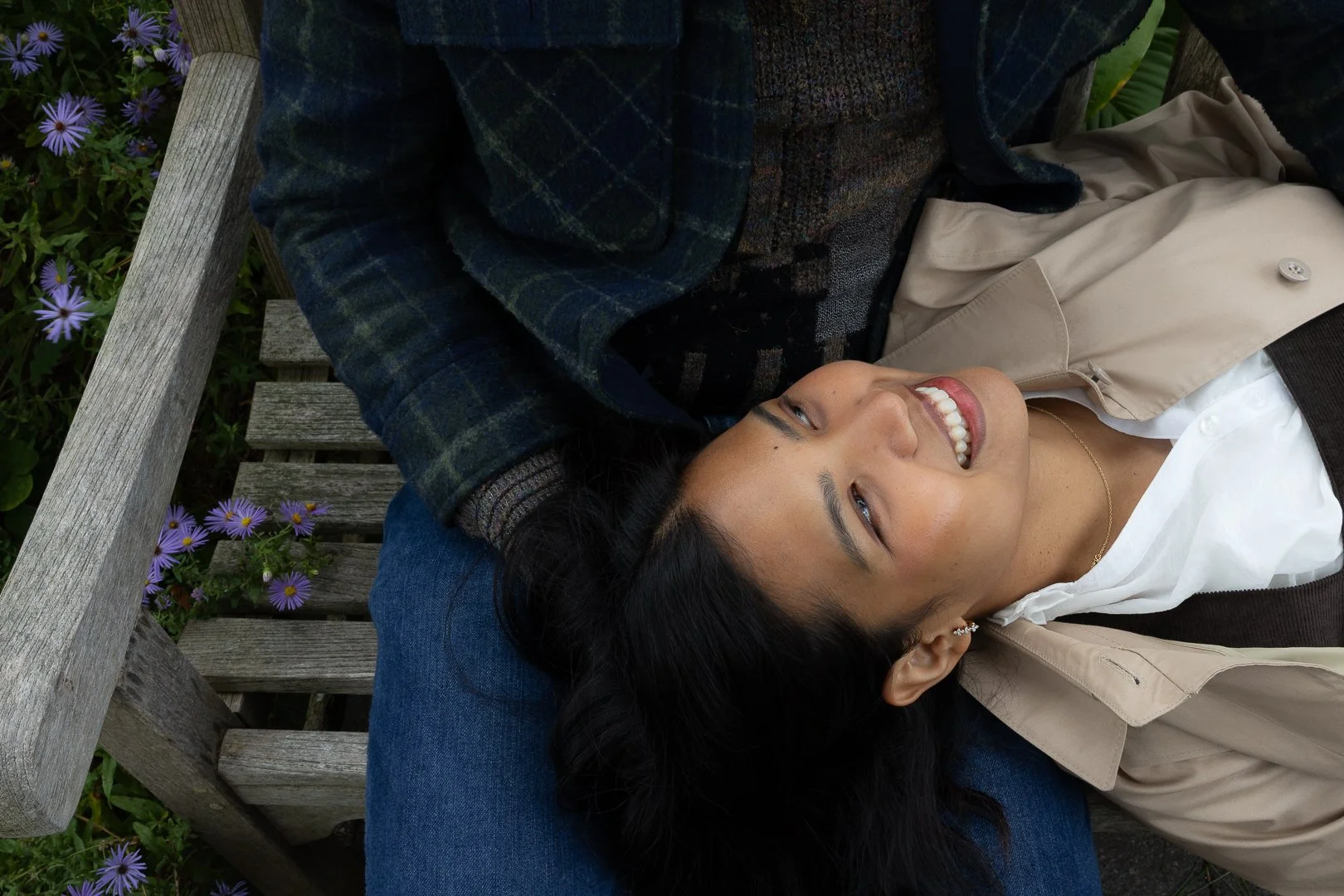 A woman lying on a park bench, smiling with her head resting on a person's leg. She is wearing a beige jacket, white shirt, and earrings, with dark hair and a necklace. Purple flowers are visible on the ground next to the bench.