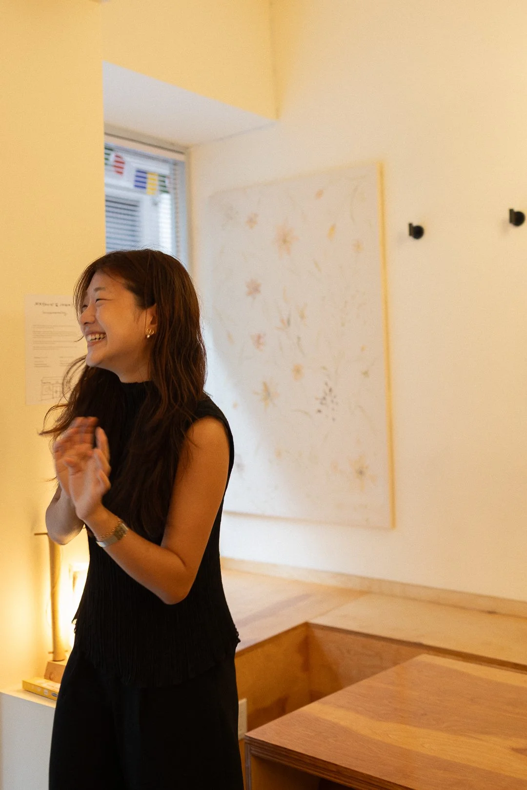 A woman with long brown hair, smiling and laughing, standing indoors in front of a light-colored wall with wall art and hooks, wearing a black sleeveless top and accessories.