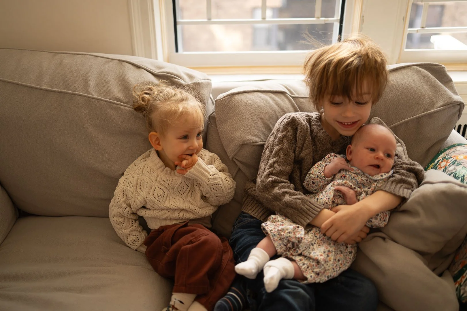 A young girl with blonde hair in a knit sweater sitting on a beige couch, watching two older siblings with a baby sibling, one holding the baby, in a cozy living room with a window in the background.
