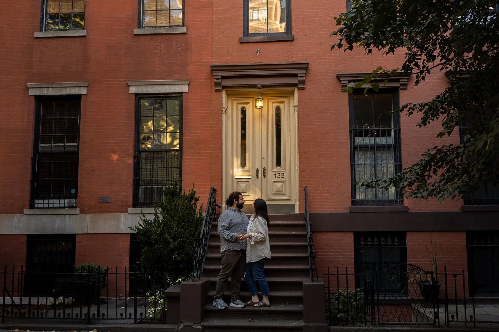A couple holding hands and looking at each other on a front porch of a brick apartment building at dusk.