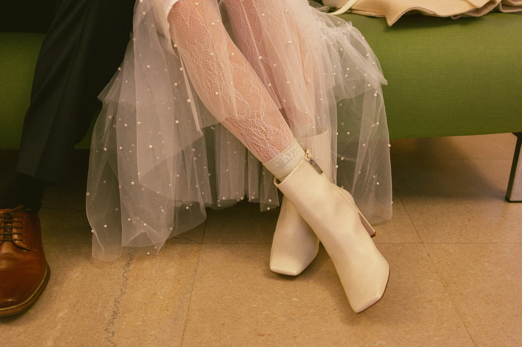 Close-up of a woman's legs, wearing pink fishnet stockings with pearl embellishments, a sheer tulle skirt with pearl accents, and a pair of white heeled ankle boots. The woman is seated on a green bench, with part of a patterned shoe and a black coat