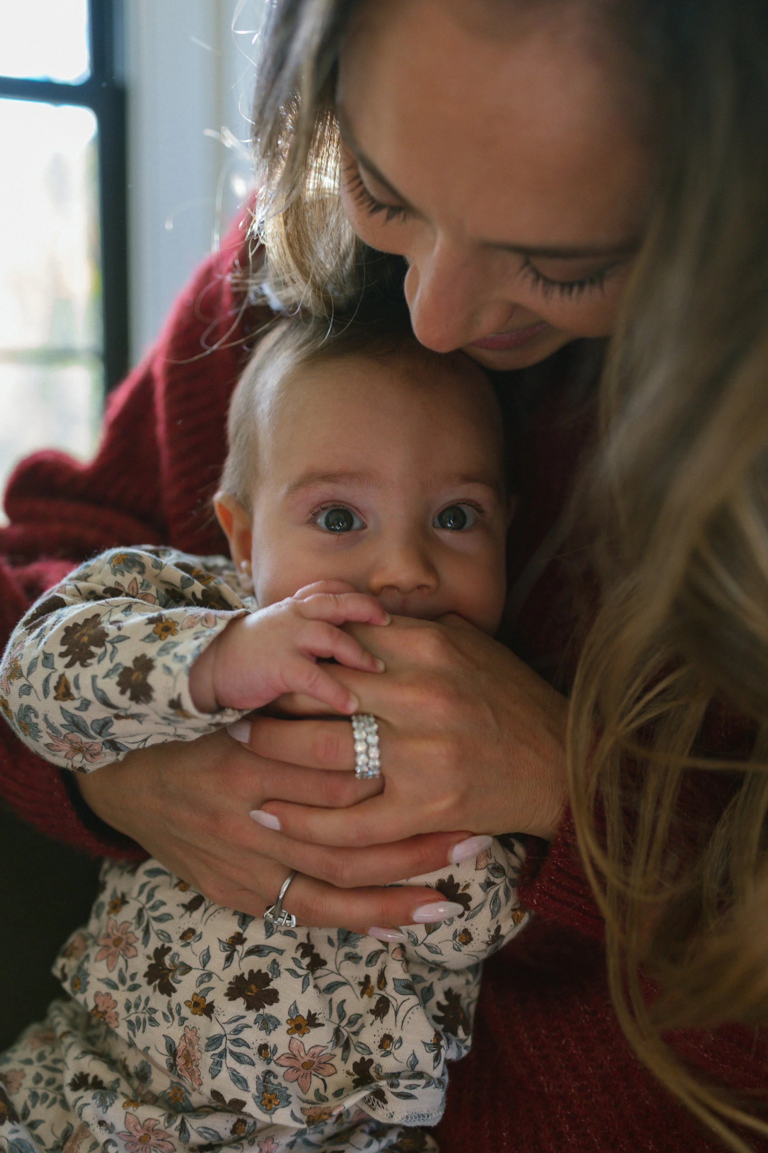 A woman holding a baby close, with the baby looking directly at the camera. The woman is leaning down, and the baby has big blue eyes, and wears a floral patterned shirt. The woman has long blonde hair and wears a red sweater and a ring with multiple