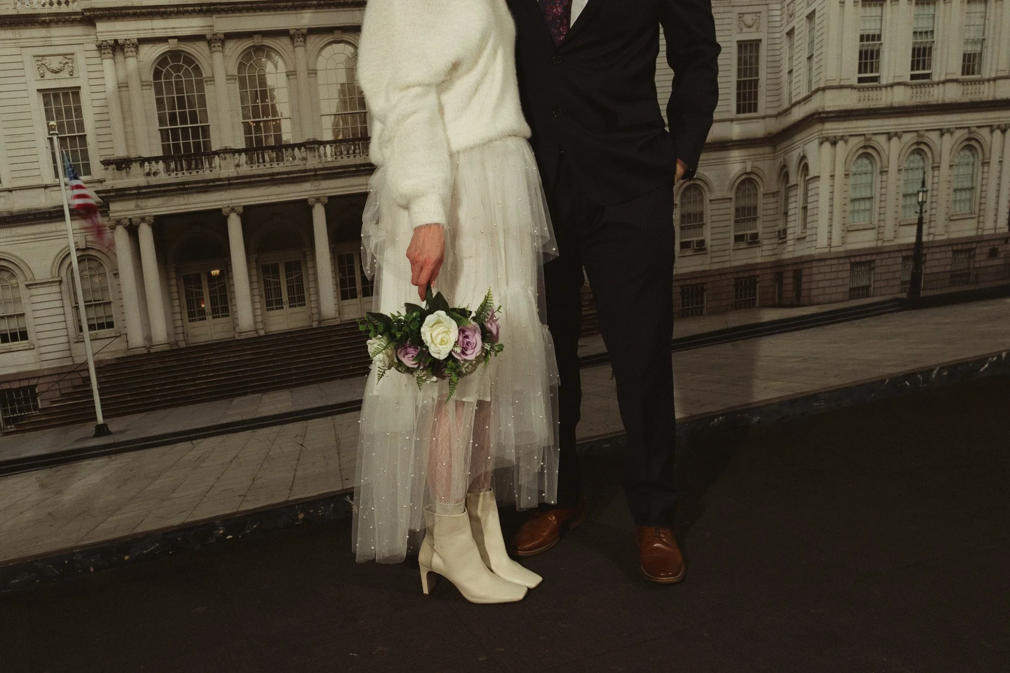 Close-up of a bride and groom standing together, holding a bouquet of flowers, with a historic government building in the background.