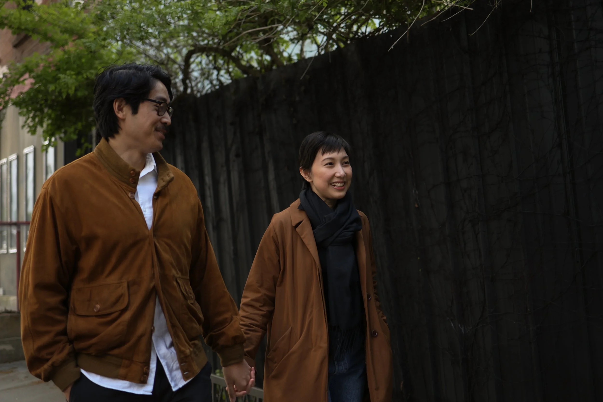 A man and woman walking together outdoors, holding hands and smiling, with trees and a black fence in the background.