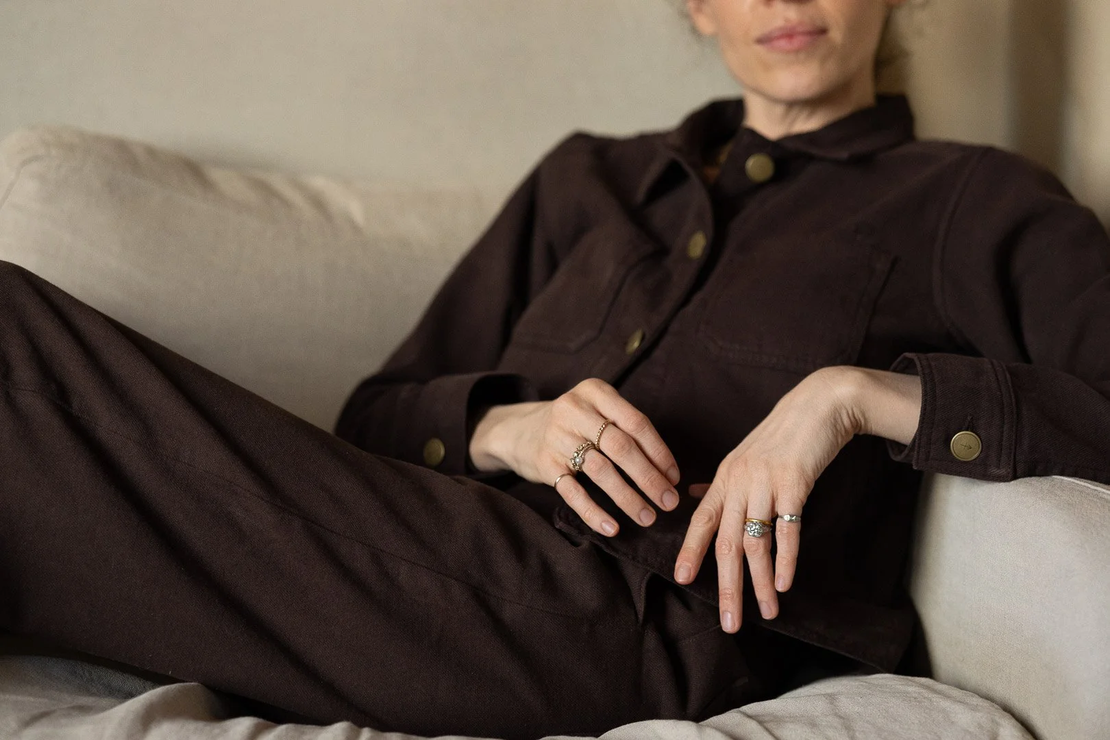 A woman wearing a dark brown jacket and matching pants, seated on a beige sofa, with jewelry on her fingers, in a relaxed pose.