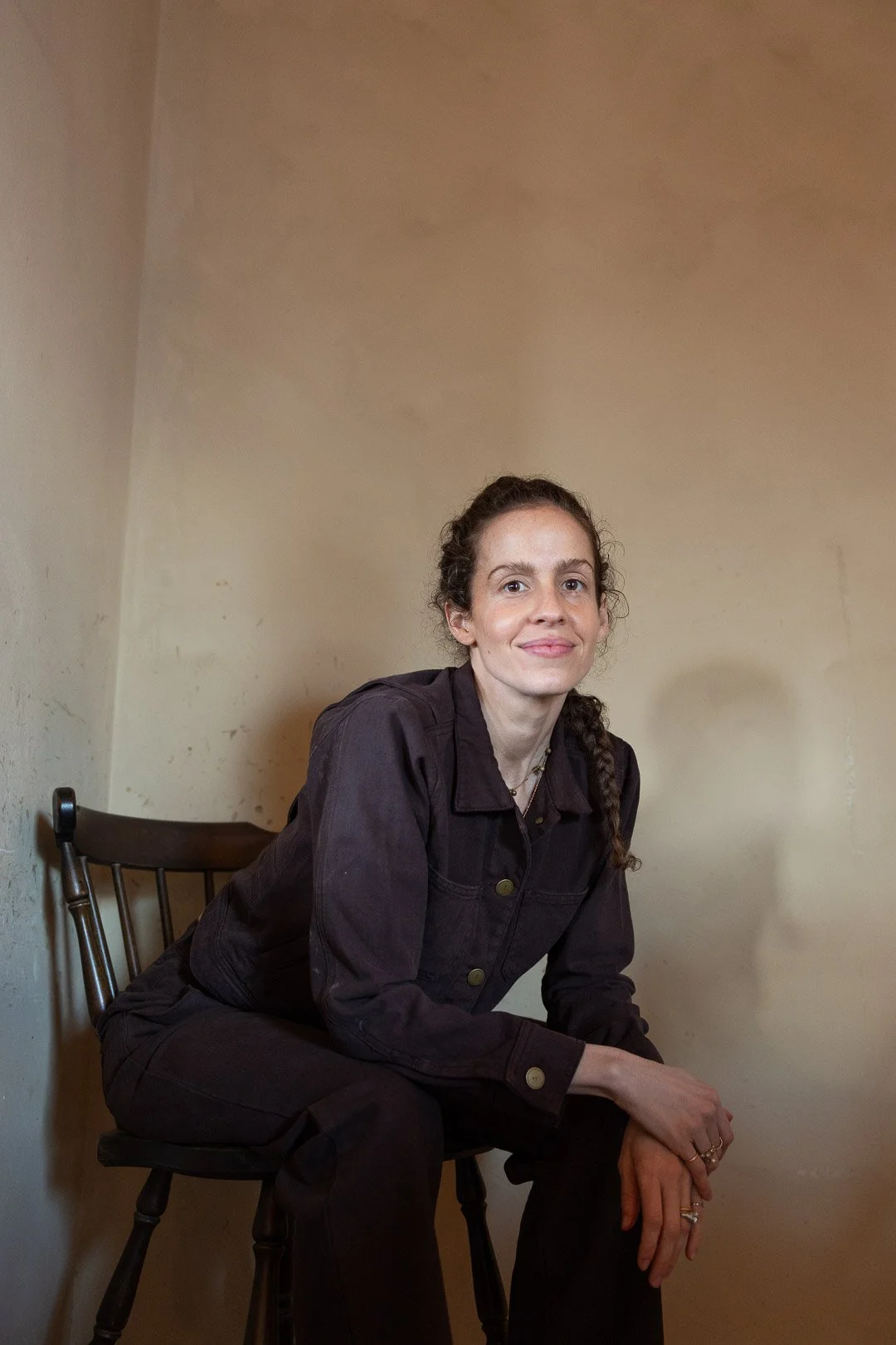 A woman with curly brown hair sitting on a wooden chair in a plain beige room.