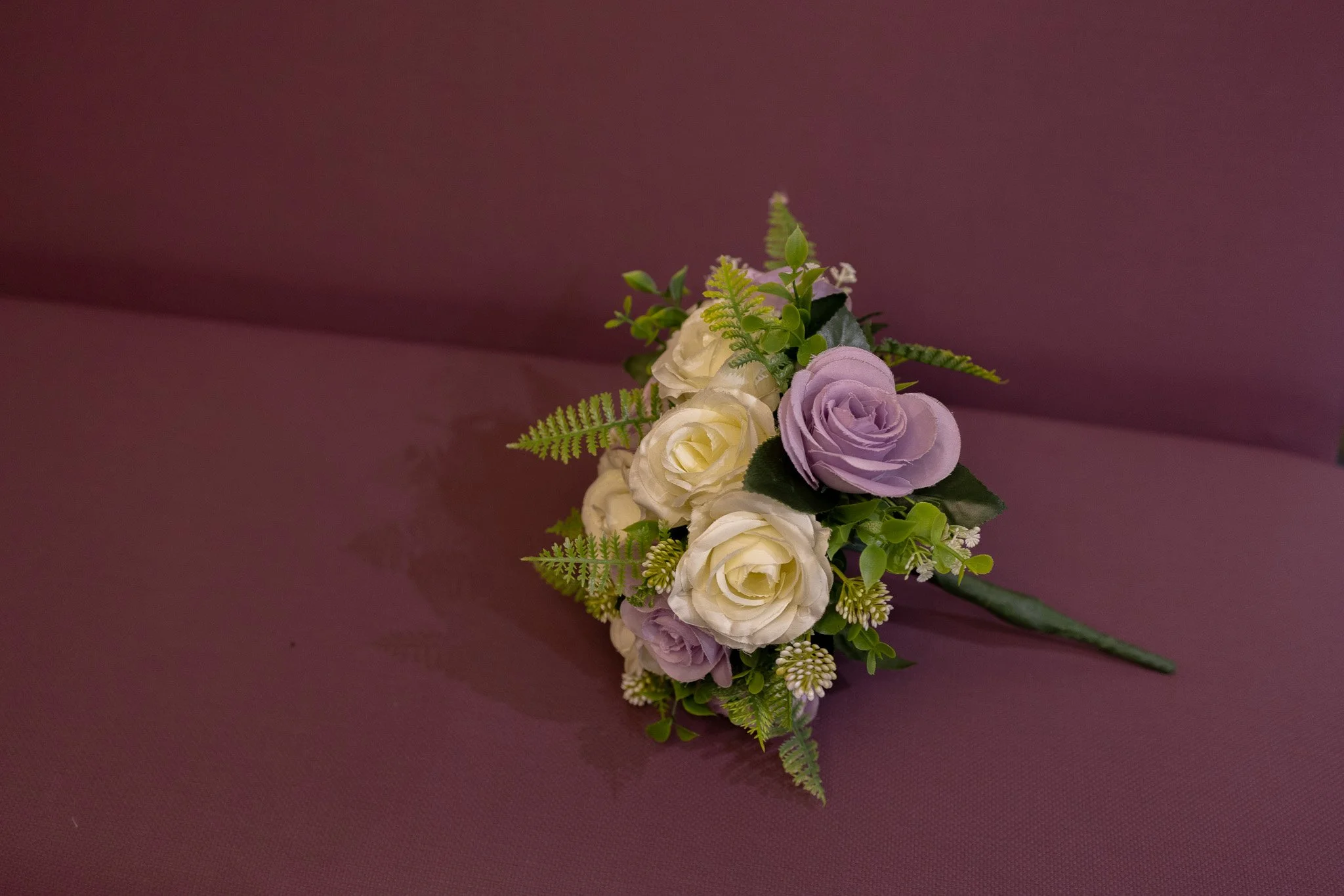 A small floral boutonniere with purple and white roses and green foliage, resting on a purple surface.