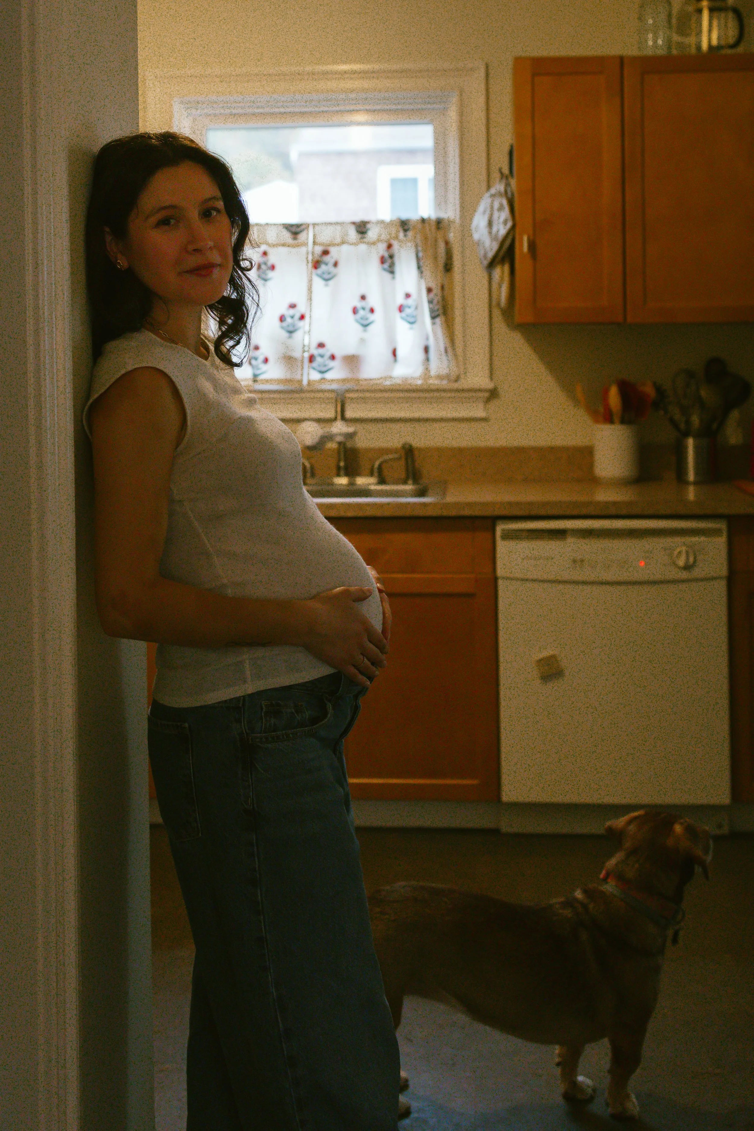 A woman standing in a kitchen with a pregnant belly, leaning against a wall, wearing a sleeveless top and jeans, with a dog nearby.