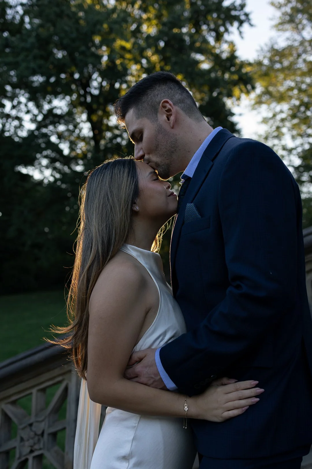 A couple in wedding attire embraces outdoors during sunset, with the man kissing the woman's forehead.