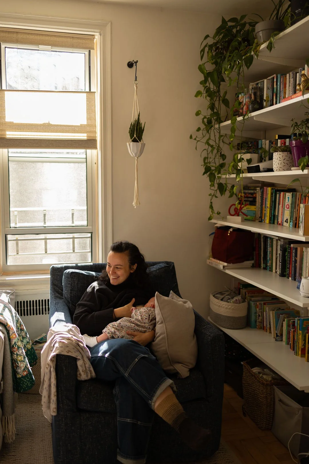 A woman sitting on a dark blue armchair holding a baby, smiling, in a cozy living room with a bookshelf, a window, a hanging plant, and various household items.
