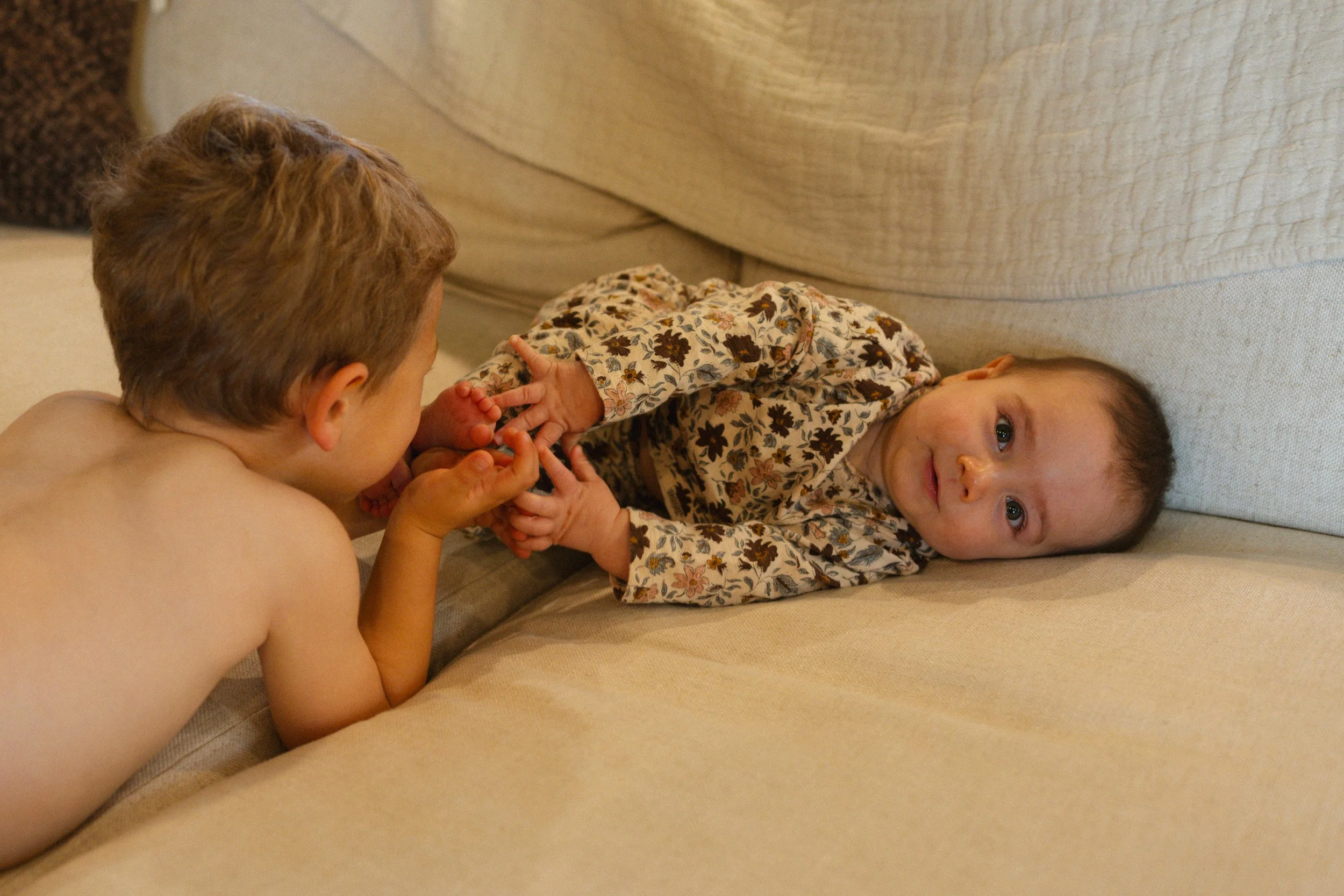 A young boy is playing with a baby girl on a beige couch. The boy, who is shirtless, is gently holding the baby's hands, and the girl is lying on her side, wearing a floral dress, looking at the camera.