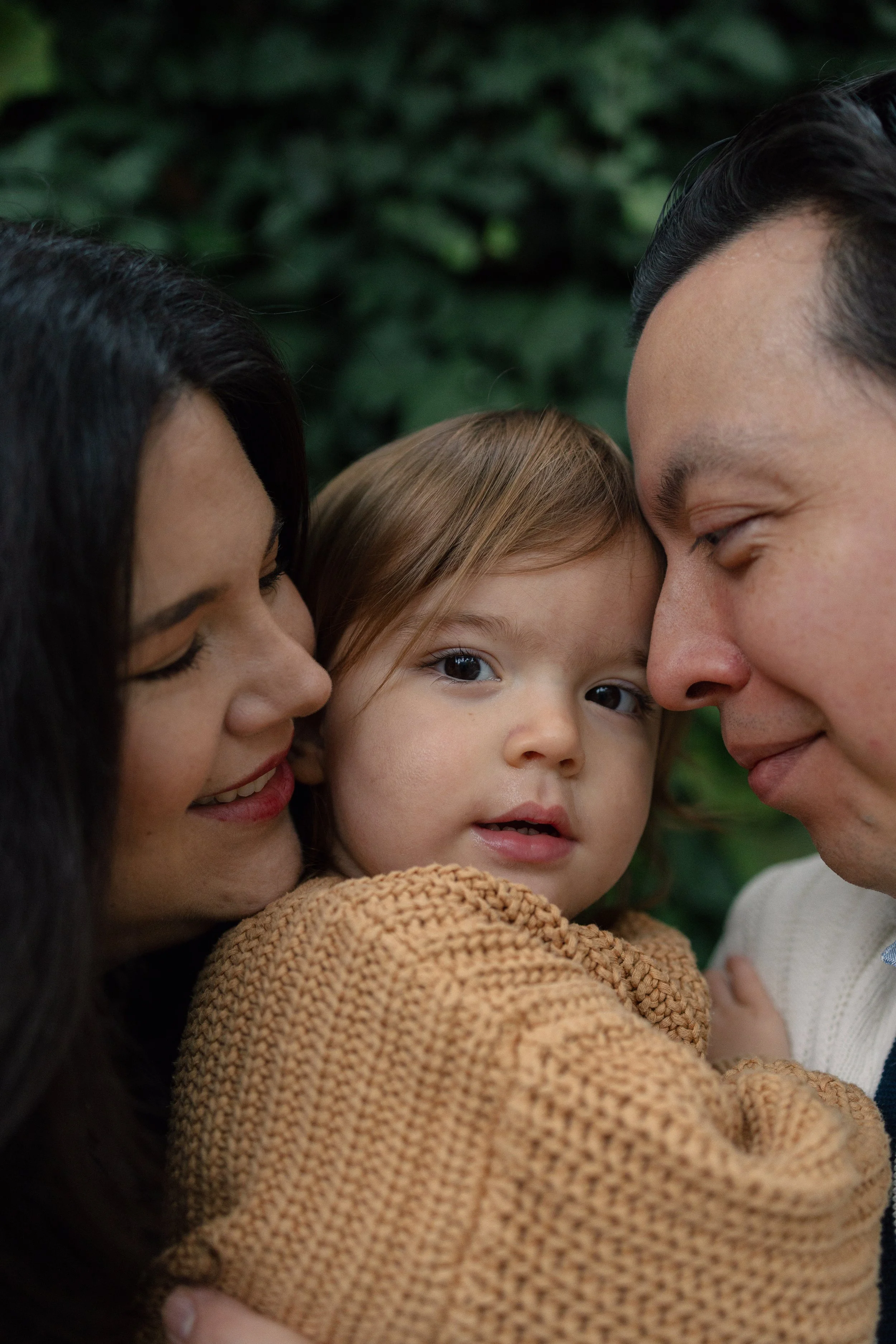 A close-up of a young girl with brown hair being embraced by a woman with dark hair on one side, and a man with dark hair on the other, both smiling gently. Their faces are close together indoors, with a dark green leafy background.