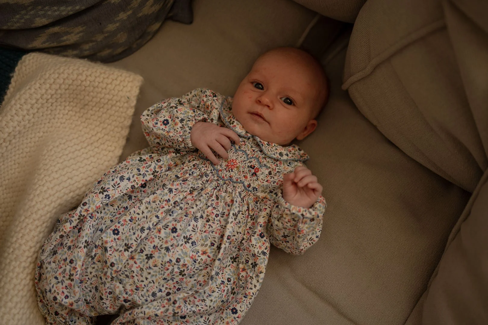A baby laying on a beige couch, wearing a floral dress, with a cream-colored knitted blanket nearby.