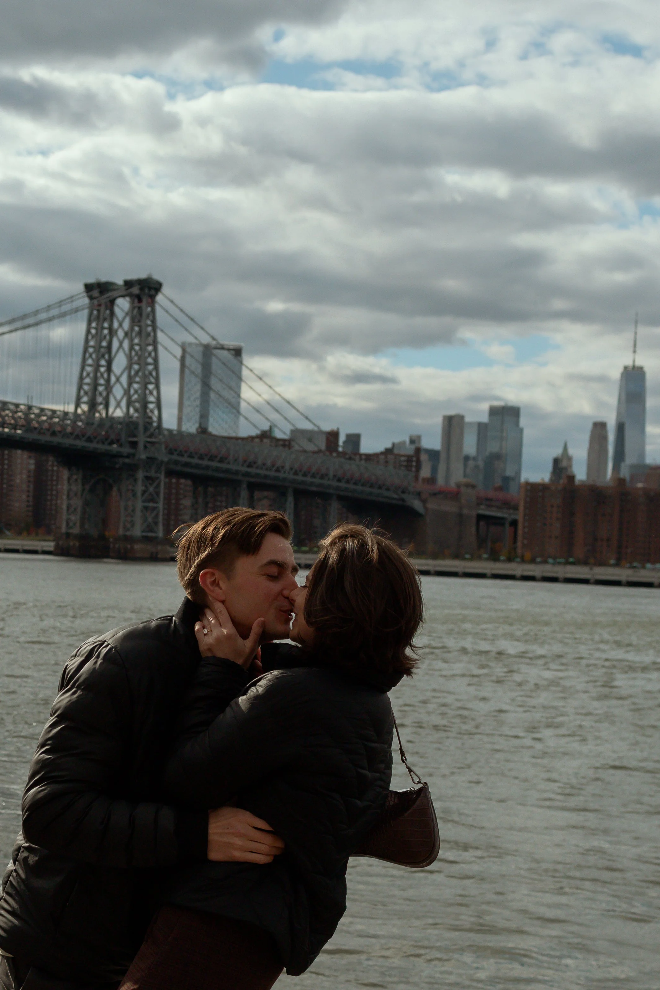 A couple embraces and kisses on the shoreline with the Manhattan Bridge and New York City skyline in the background on a cloudy day.
