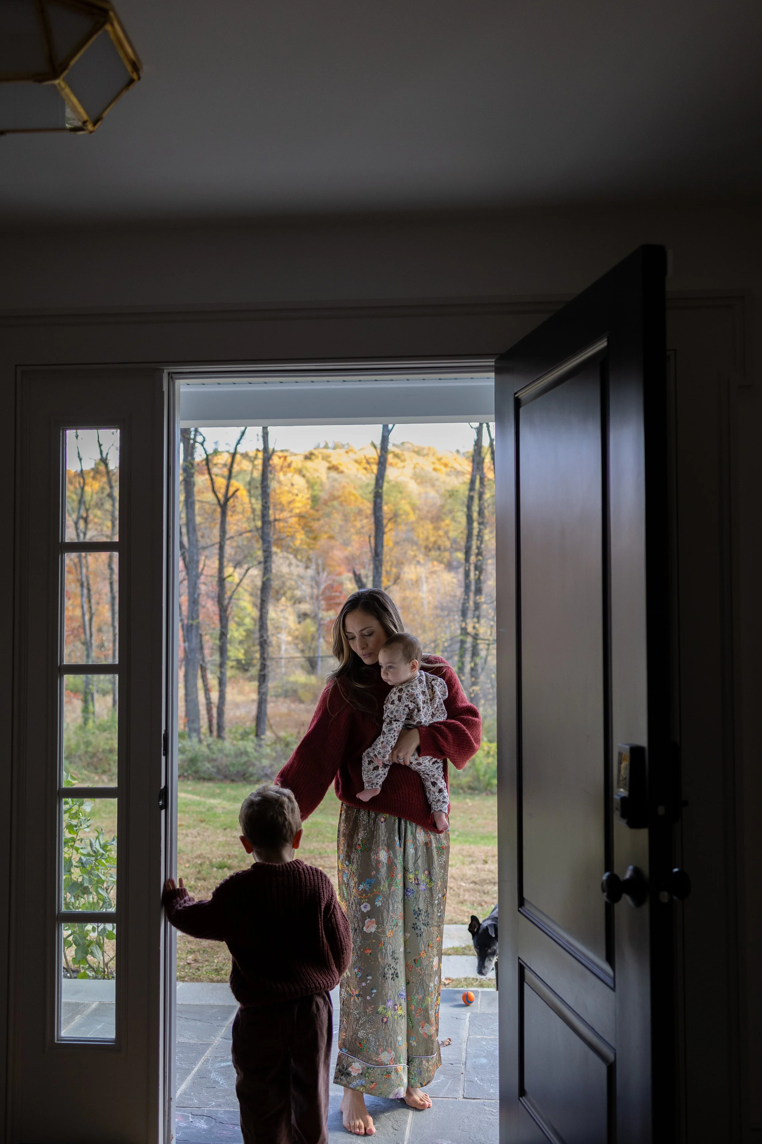 A woman with long brown hair wearing a red sweater and floral pants stands at the door with two small children, one with hair covered by a hood, during daytime fall, with trees with yellow, orange, and green leaves in the background, and a dog outsid