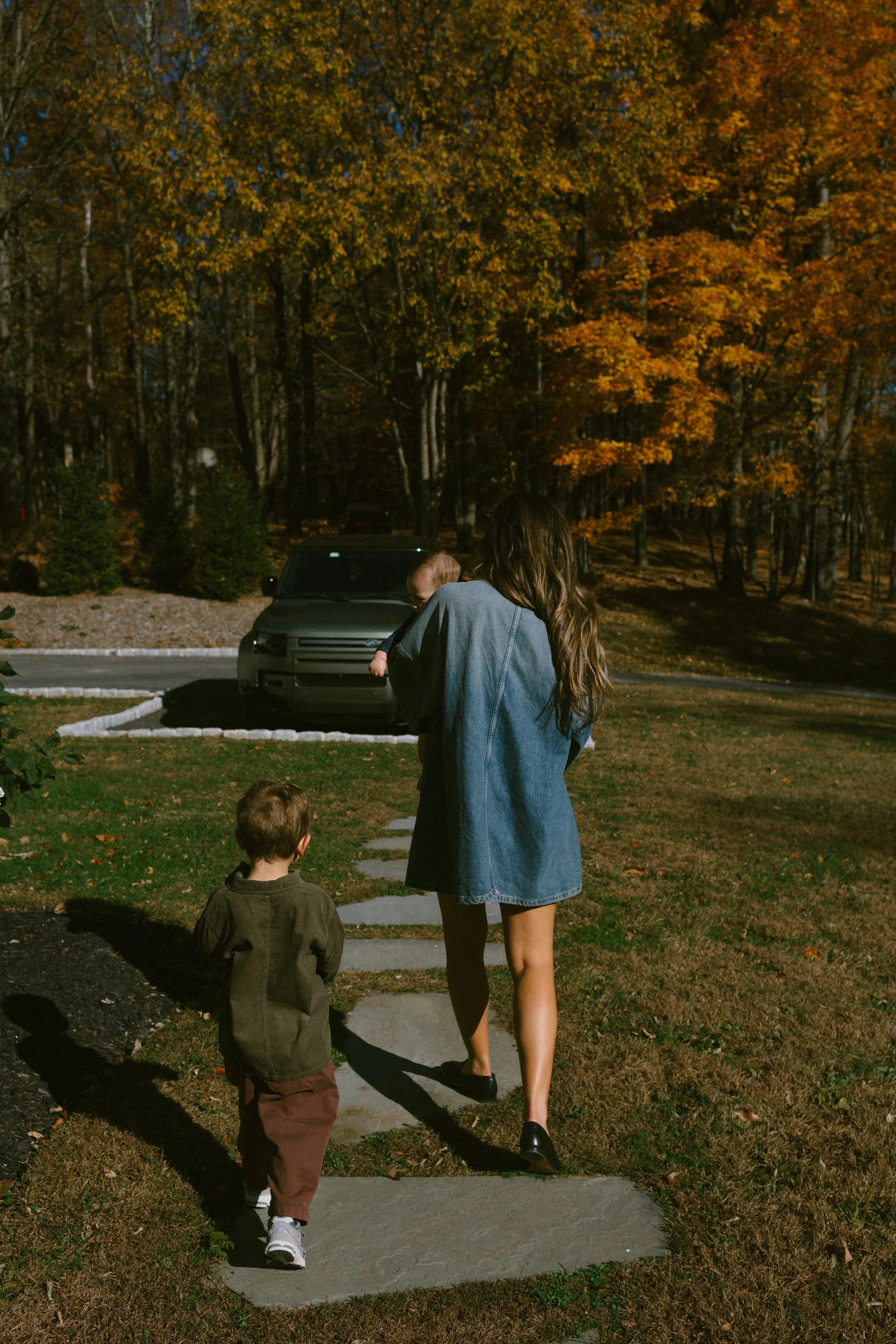 A woman walking on a stone pathway with two young children in a park with trees displaying autumn foliage.